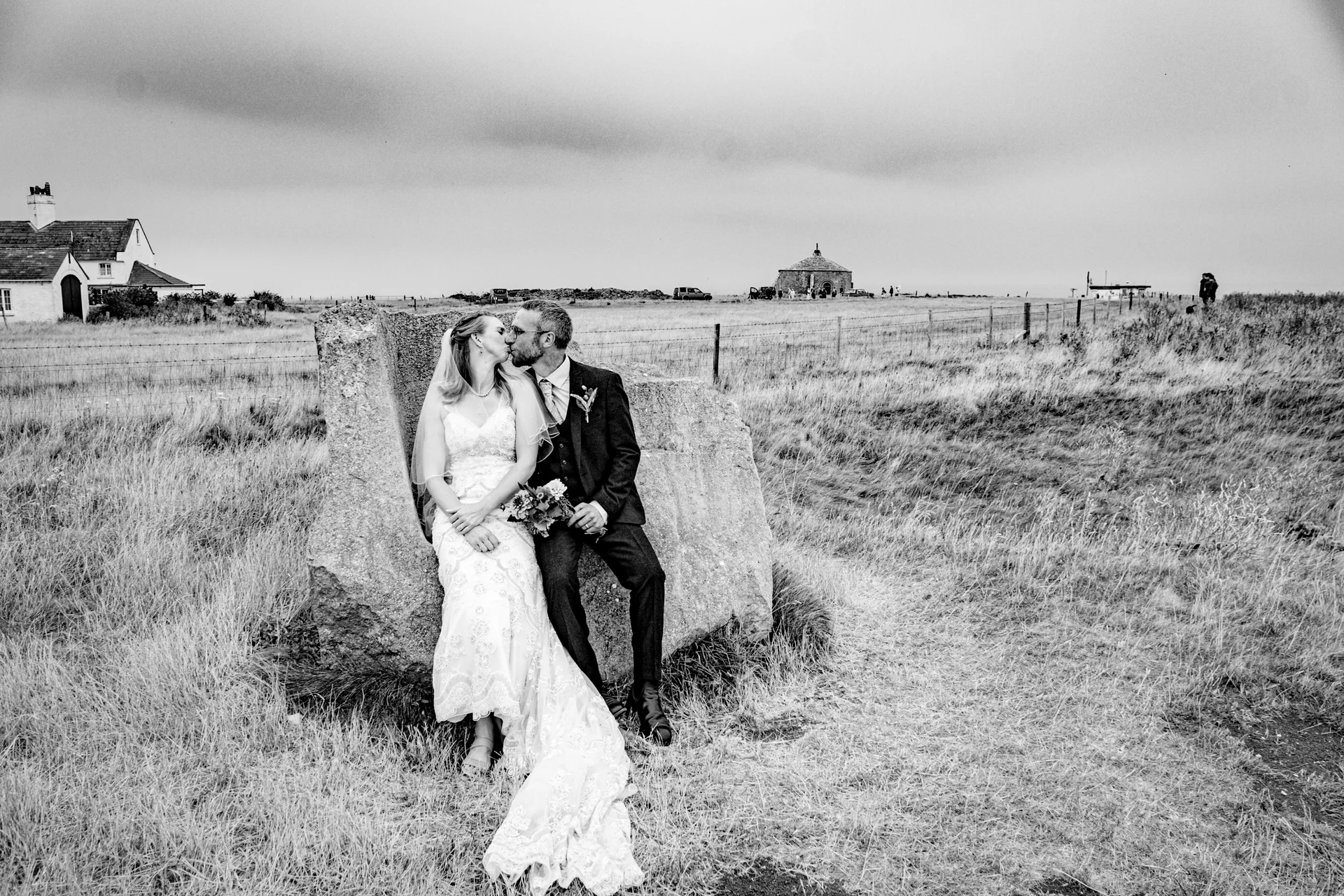 A newlywed couple shares a tender kiss while sitting on a large weathered rock amidst an expansive, moody moorland landscape under an overcast sky.
