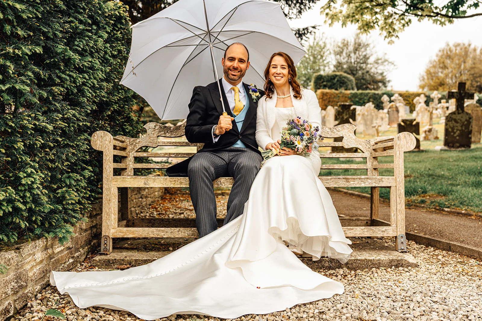A smiling bride and groom sit together on a wooden garden bench outdoors holding a white umbrella, with the bride displaying her long satin train and colourful bouquet.