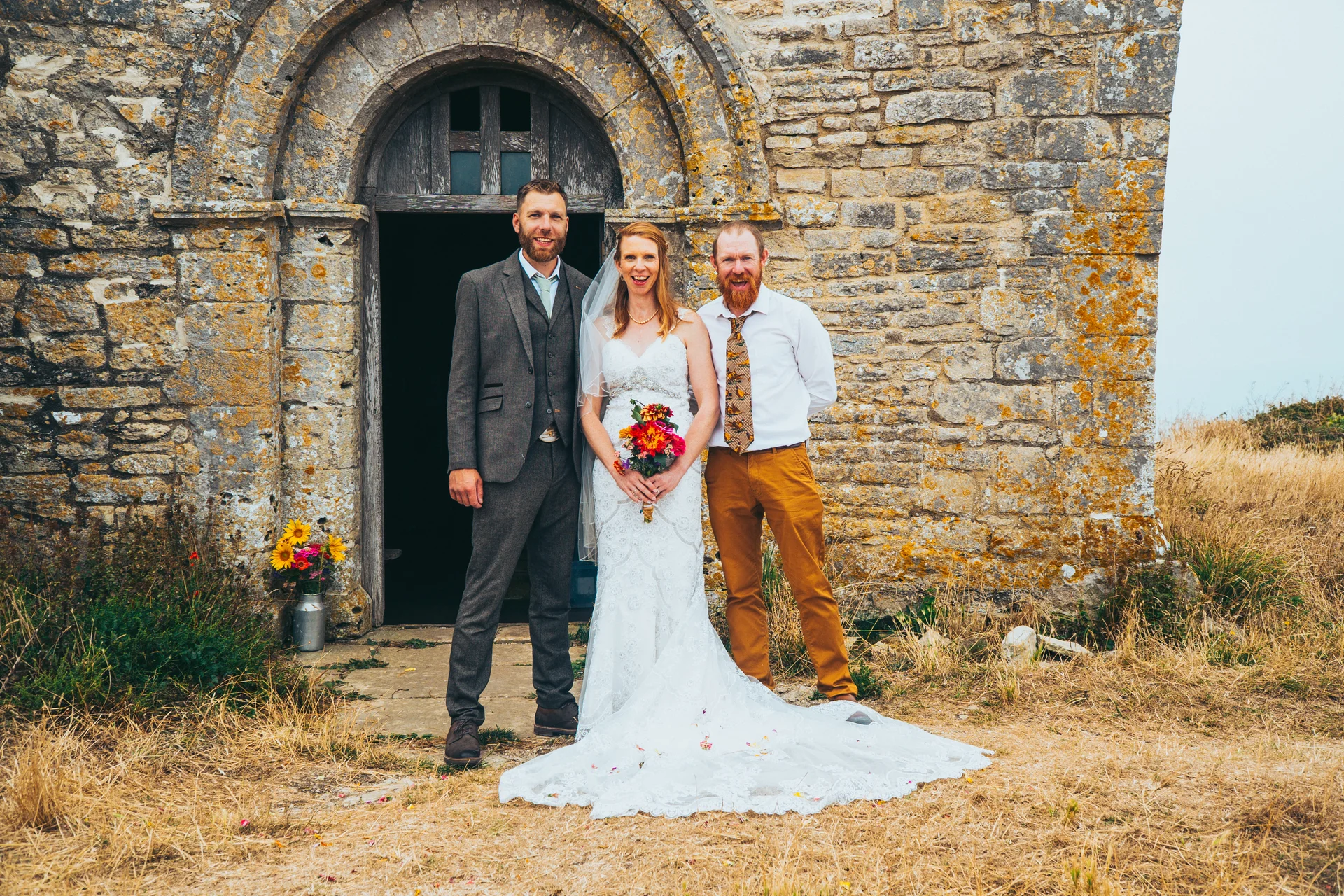 A smiling bride holding a colourful bouquet stands proudly with the groom in a tweed suit and another male wedding party member outside the arched wooden doorway of a rustic stone church.
