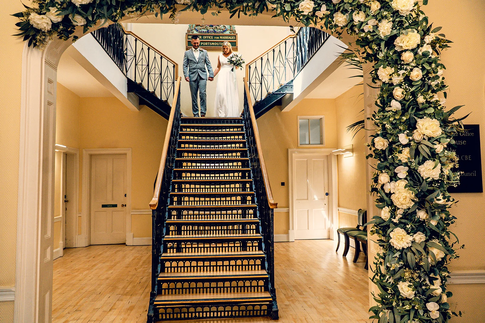 A newlywed couple holding hands descends a grand staircase framed by an opulent white and green floral arch inside a registry office.