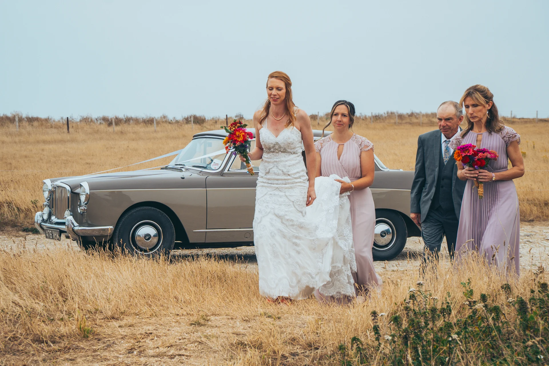 The bride holding a vibrant bouquet walks through a dry field alongside her bridesmaids and father, with a classic vintage car parked in the background under an overcast sky.