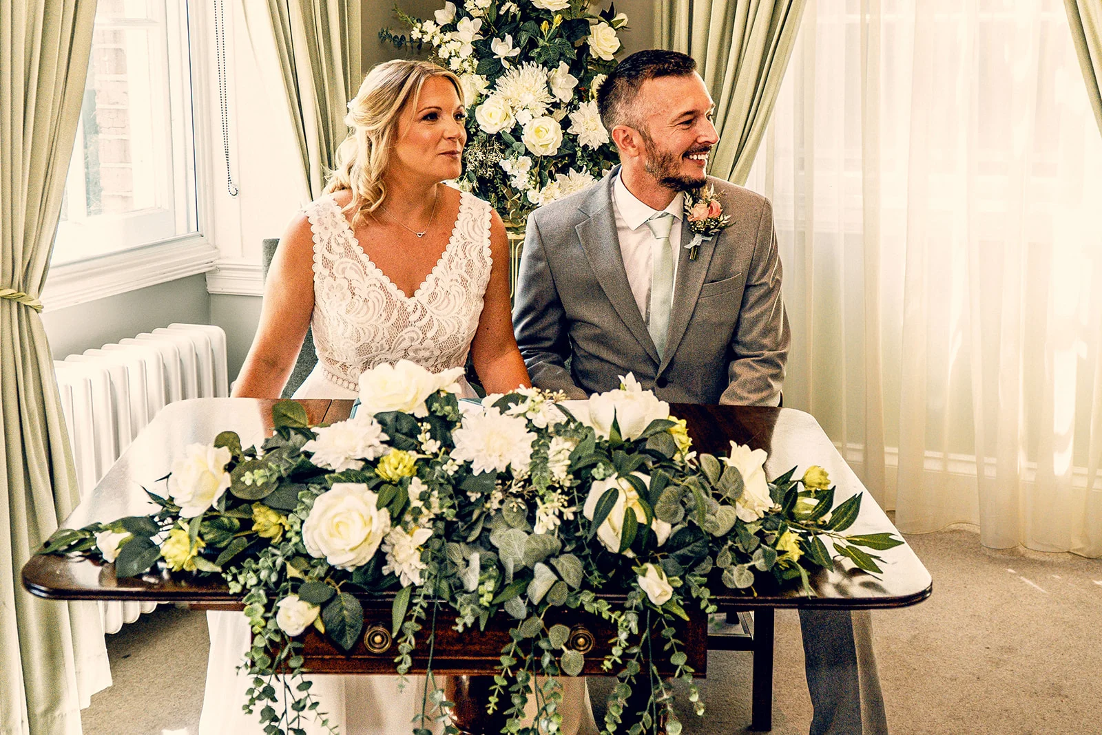 A smiling bride in a lace dress and groom in a grey suit sit together at a table adorned with lush white floral arrangements during their civil ceremony.