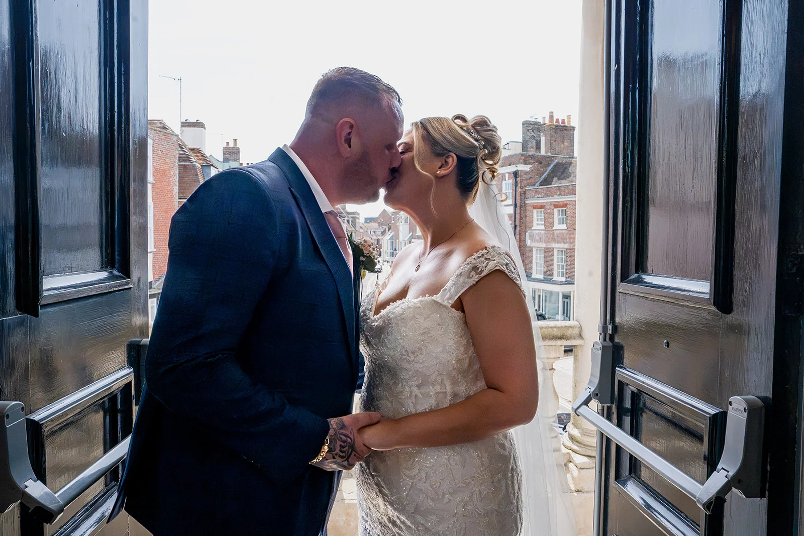 Newlywed bride and groom share a tender kiss framed by dark double doors overlooking a historic city street.