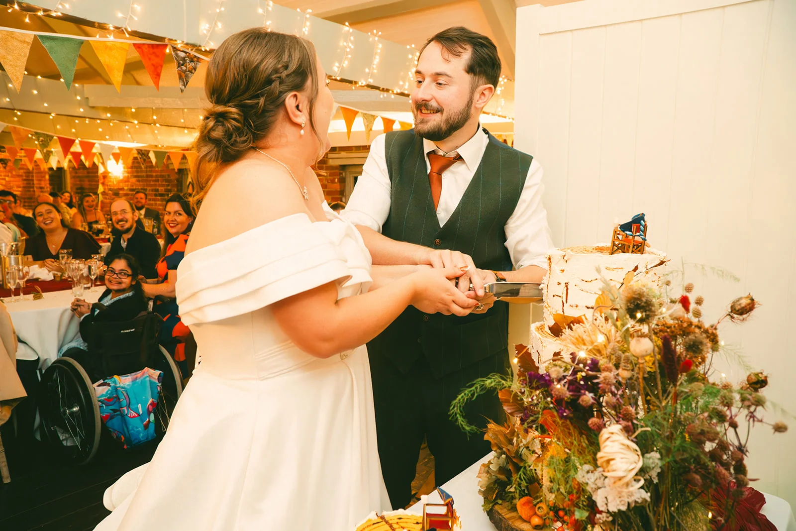 A smiling bride in an off-the-shoulder white satin gown and groom in a dark waistcoat cut their tiered wedding cake together while guests watch happily in the background.