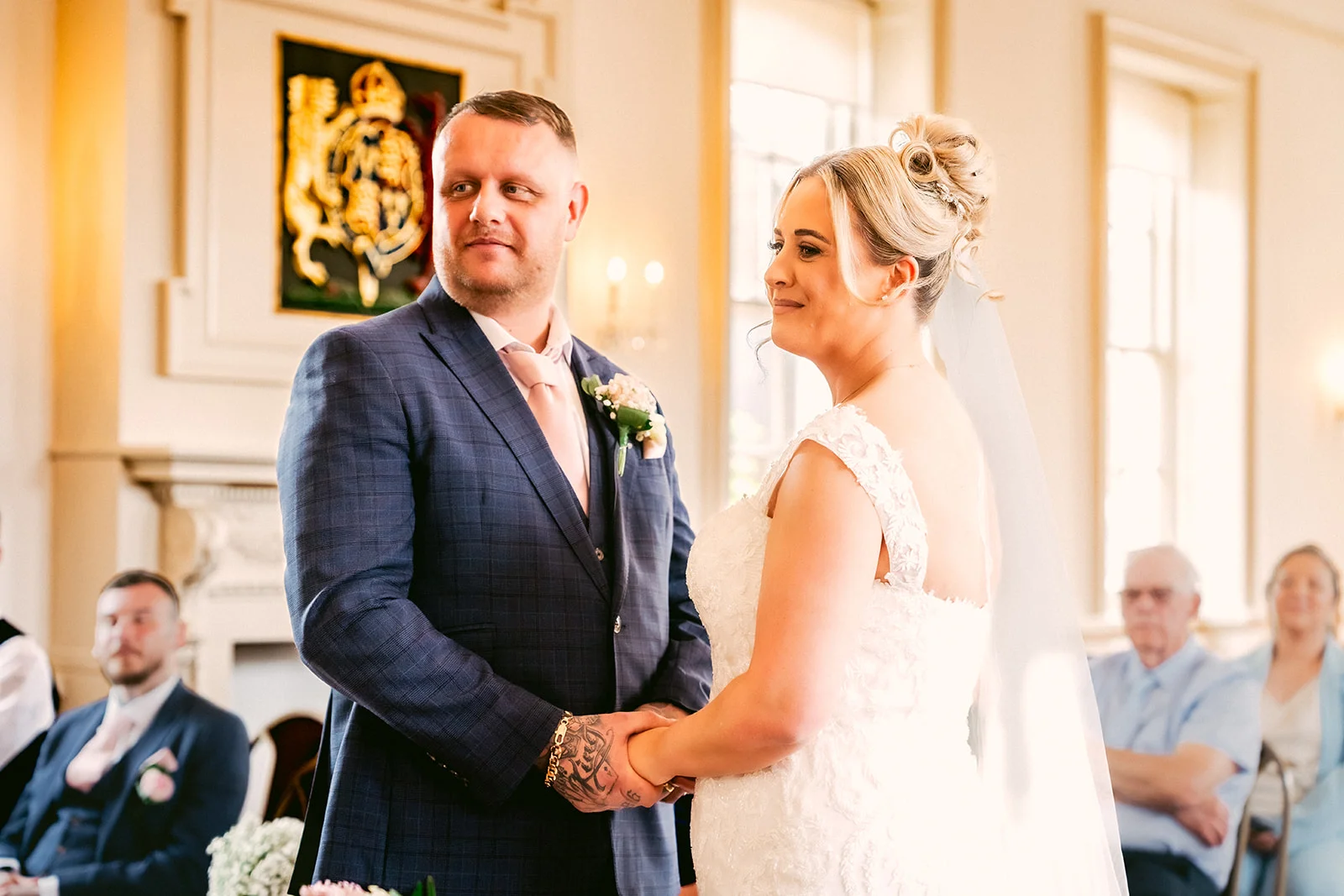 A groom in a blue checked suit and a bride in a lace wedding dress stand together holding hands during their civil ceremony.