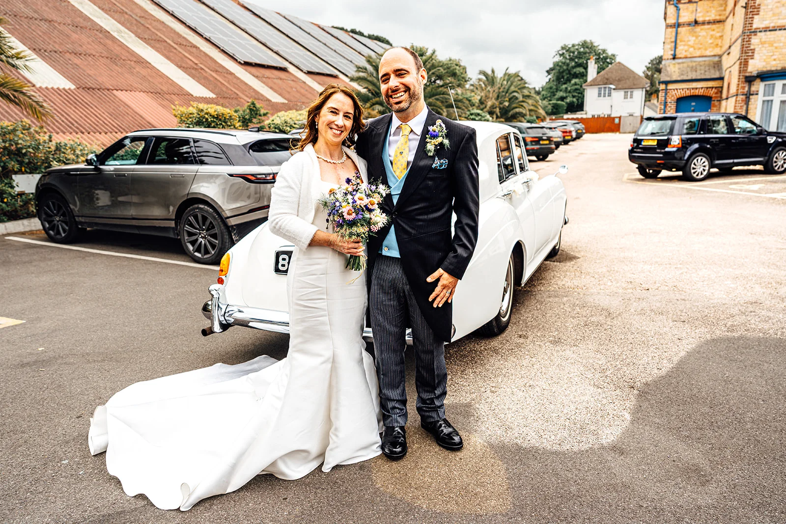 A smiling bride and groom pose happily together in a car park next to a vintage white wedding car, with the bride holding a colourful wildflower bouquet.