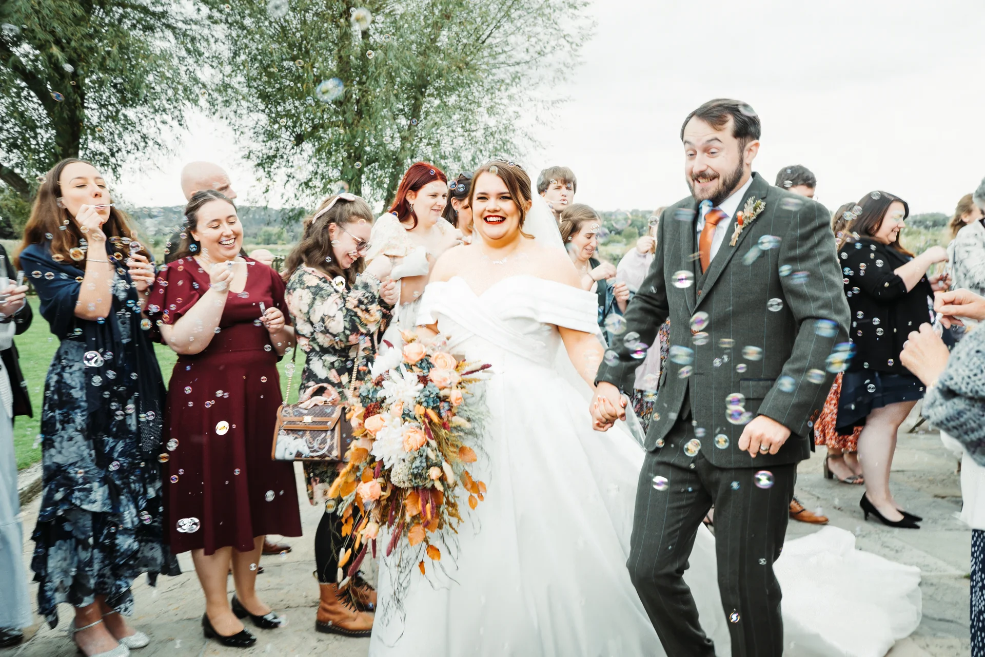A joyful newlywed couple walks hand in hand through a cascade of floating bubbles, surrounded by smiling guests blowing bubbles outdoors.
