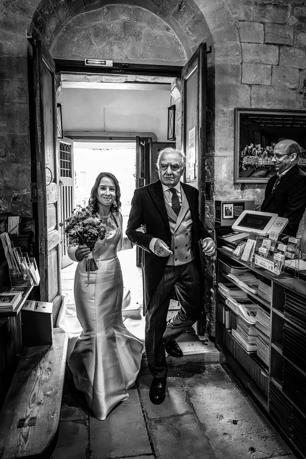 A mature bride holding a hand tied bouquet enters the church arm in arm with her father through a stone archway doorway.