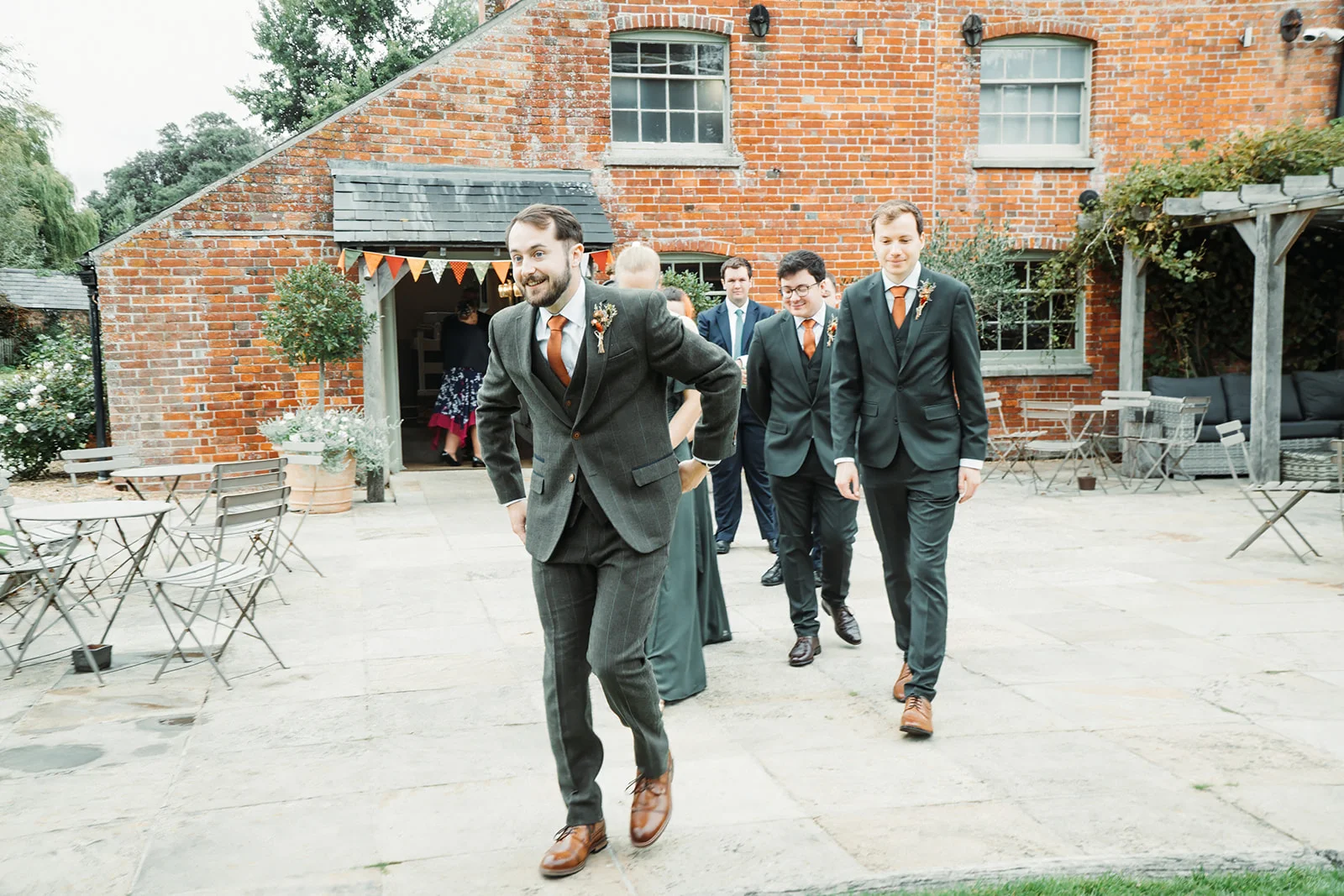 A groom and his groomsmen in tweed suits walk across a paved courtyard outside a red brick barn.