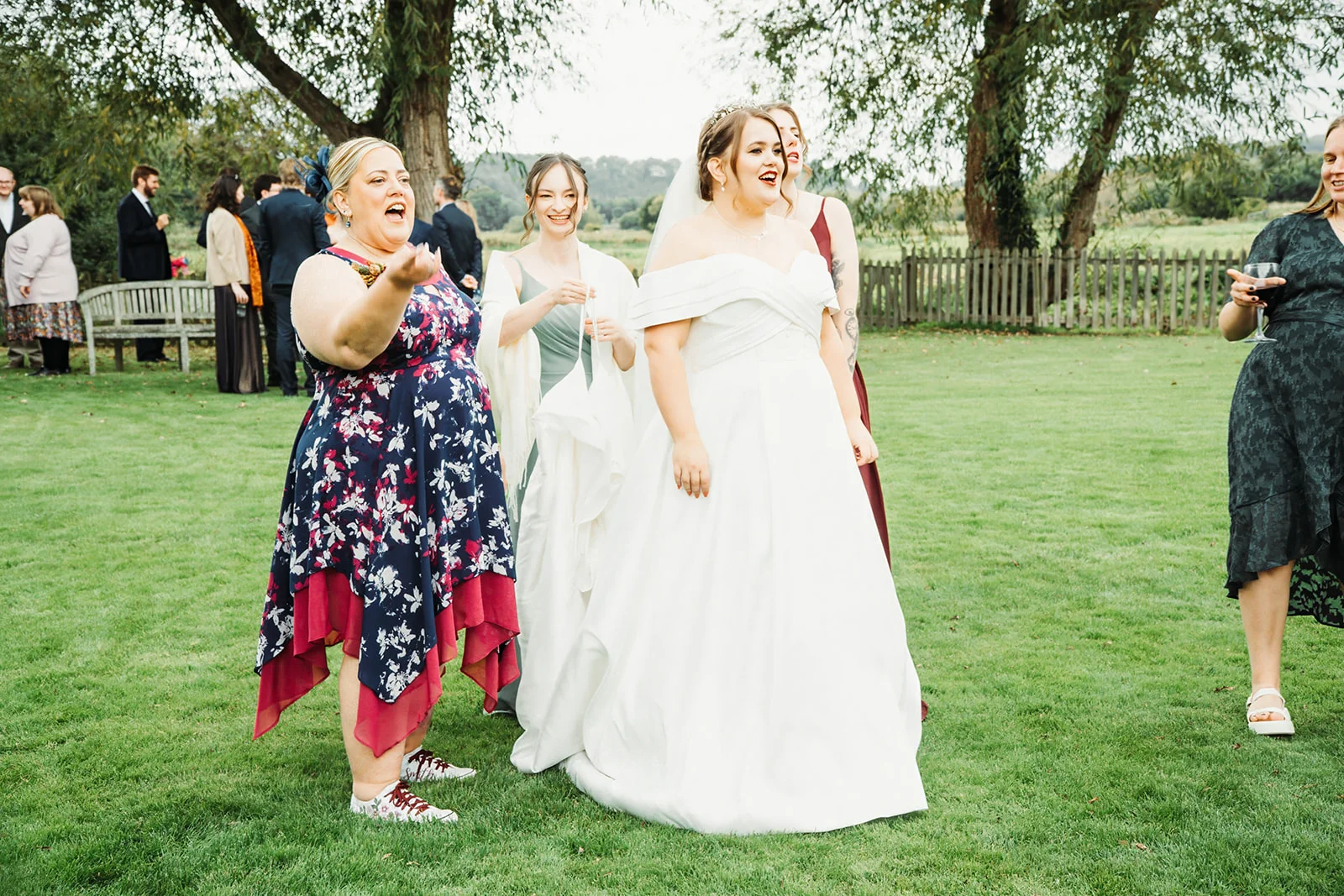 A joyful bride in a white off-the-shoulder gown stands on a green lawn surrounded by laughing guests and bridesmaids outdoors.