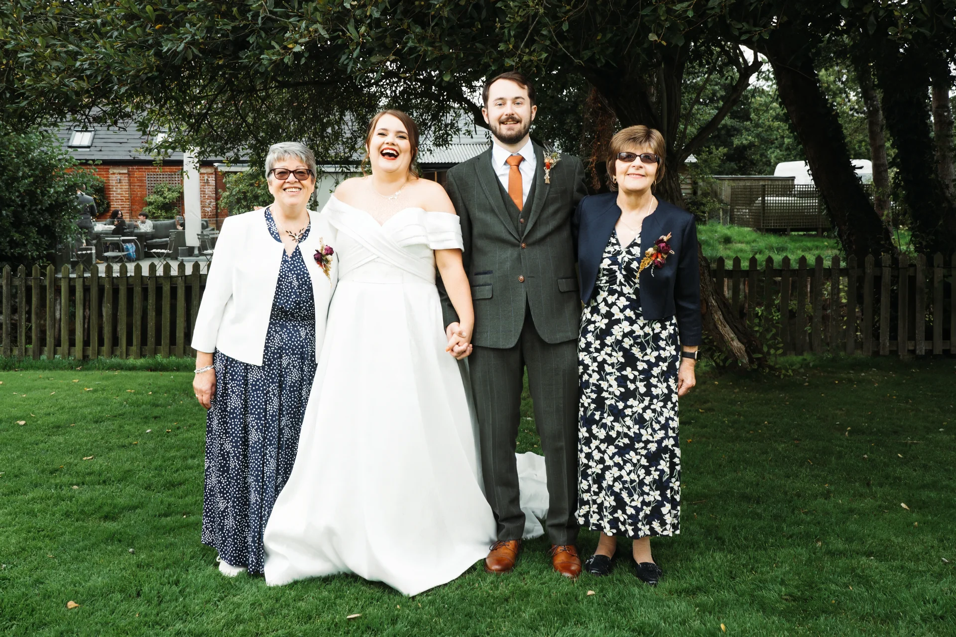 A joyful newlywed couple poses outdoors on a lush green lawn alongside two older family members, laughing happily beneath the shade of leafy trees.