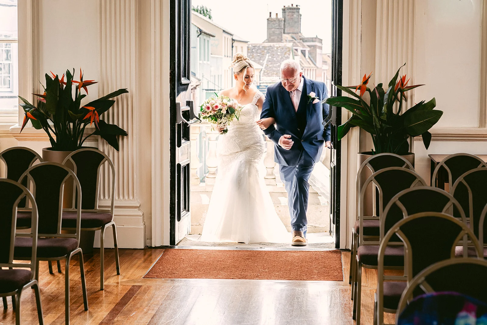 The bride enters the ceremony room on the arm of her father, framed by double doors at the top of the aisle.