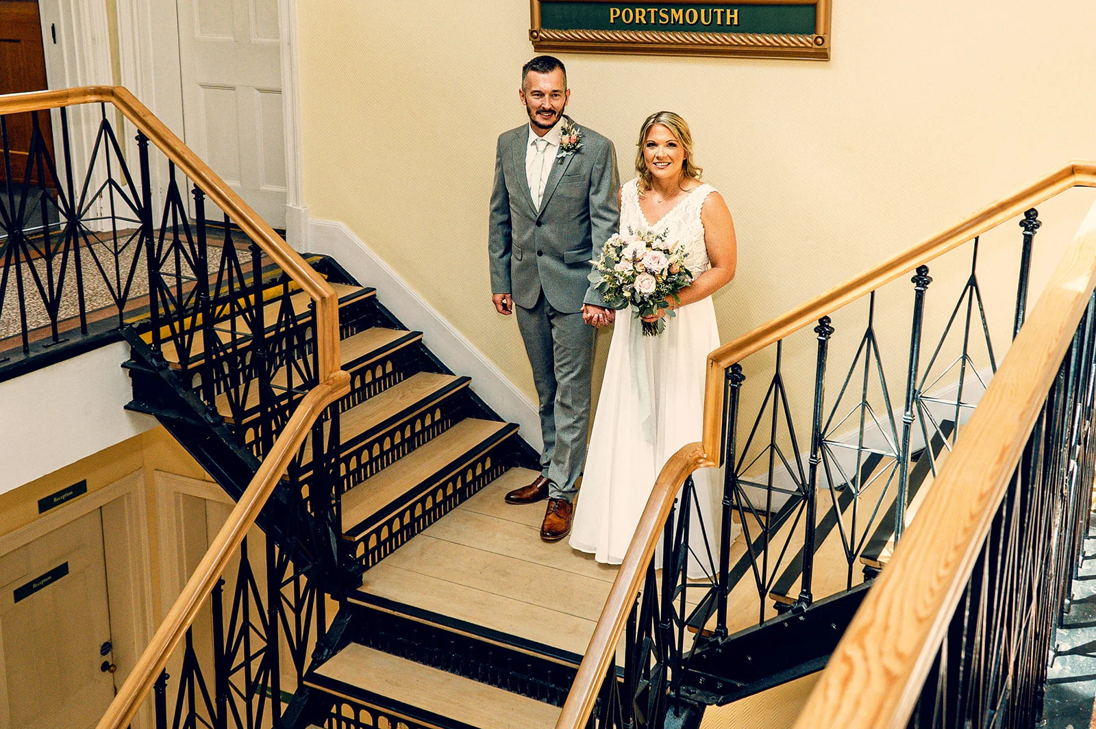 A newlywed couple smiling while standing together on a grand staircase inside a town hall, with the bride holding a delicate floral bouquet.