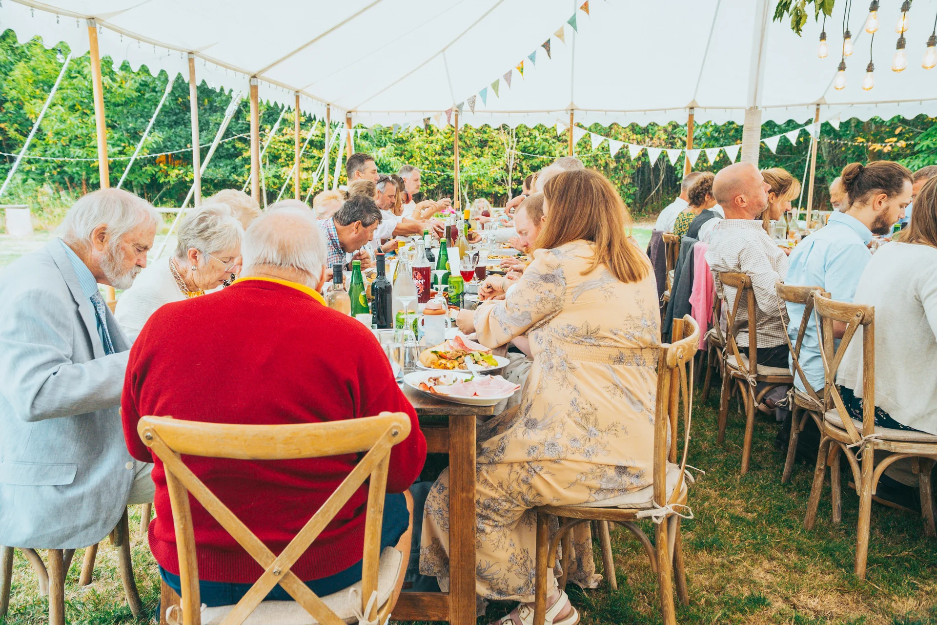 Wedding guests enjoy a relaxed wedding breakfast seated along long banquet tables inside a bright marquee decorated with bunting.