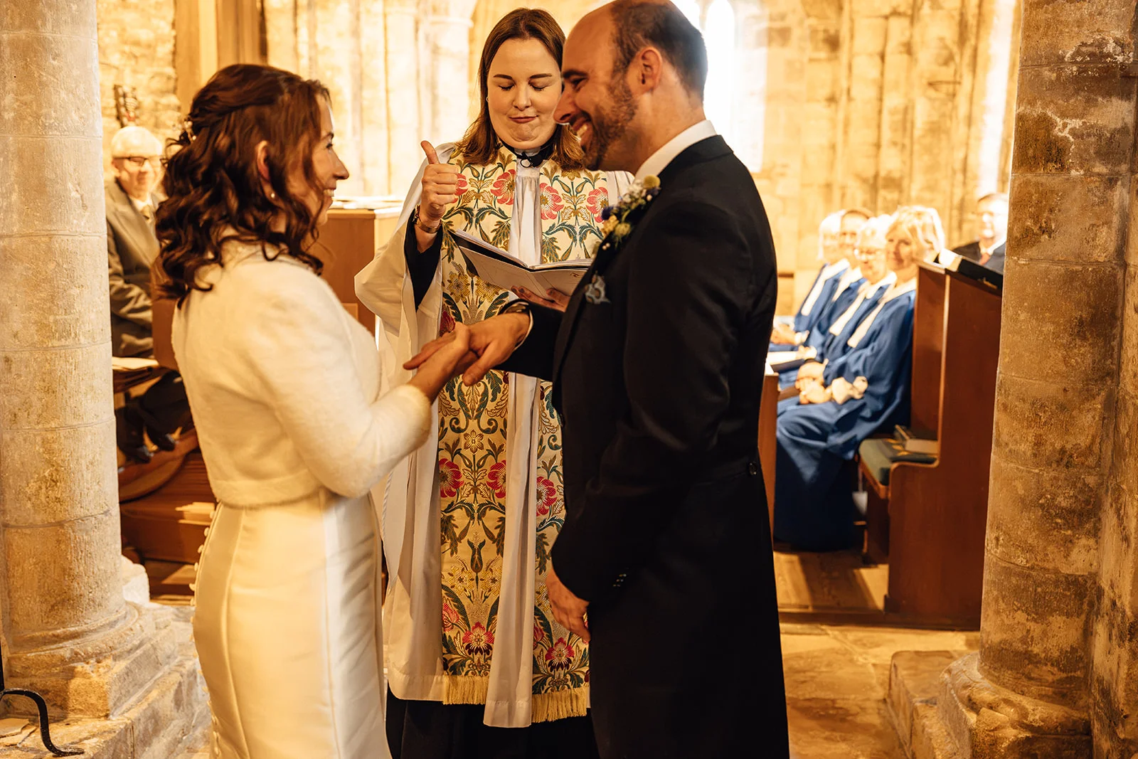A bride and groom exchange rings during their church wedding ceremony, smiling as an officiant in clerical robes looks on.