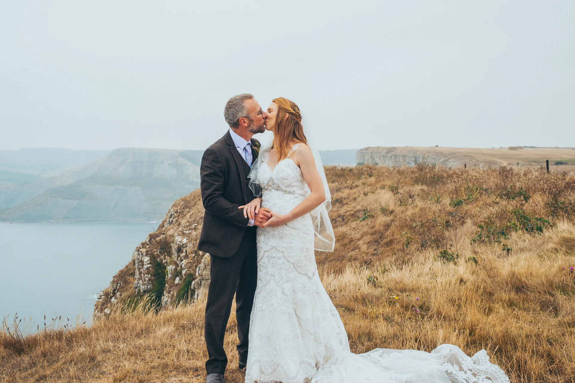 An older bride and groom share a tender kiss on a dramatic clifftop overlooking the sea on their wedding day.