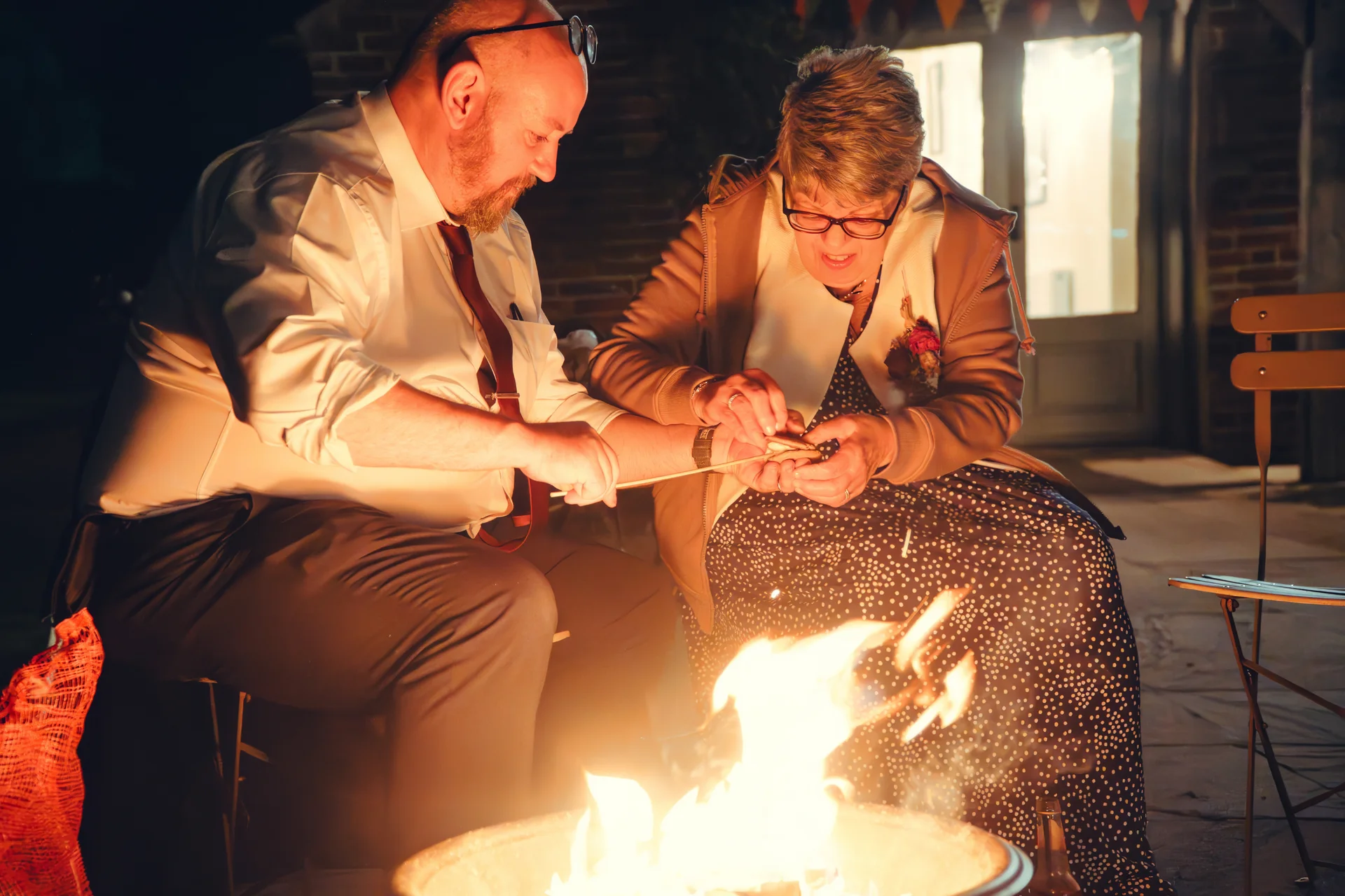 An older wedding guest couple sits close together near a glowing fire pit at night, sharing a lighthearted moment while toasting marshmallows.