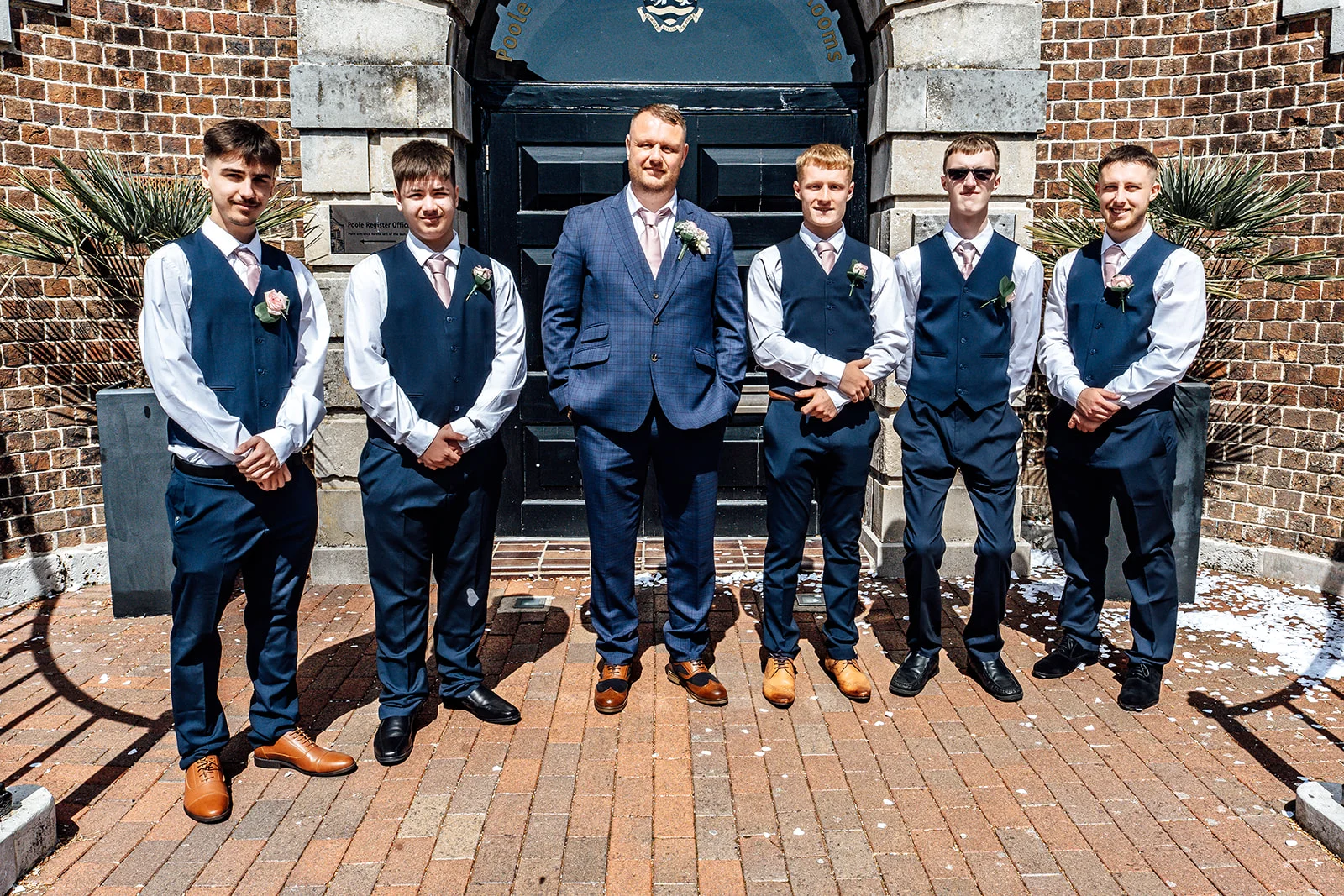 The groom and his five groomsmen stand proudly outside a brick registry office in matching blue waistcoats, pink ties, and buttonholes on a sunny day.