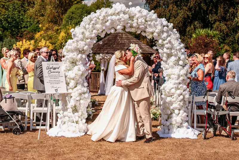 A newlywed couple shares a romantic kiss underneath a magnificent white floral arch during an outdoor garden wedding ceremony while guests watch from seated rows.