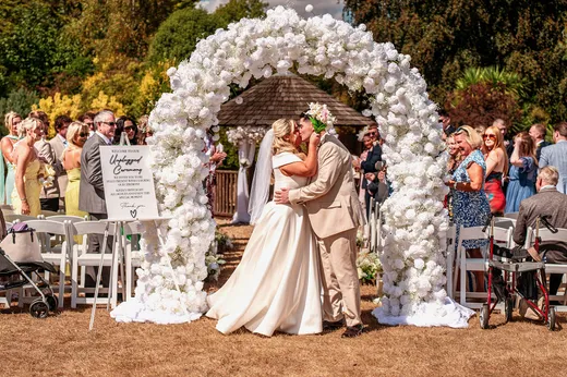 A newlywed couple shares a romantic kiss underneath a magnificent white floral arch during an outdoor garden wedding ceremony while guests watch from seated rows.