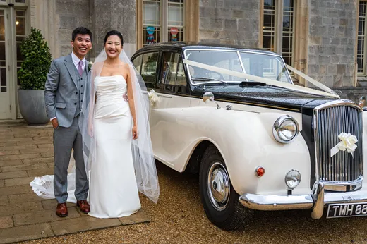A smiling bride and groom pose happily next to a vintage black and white wedding car parked outside a stone manor house.