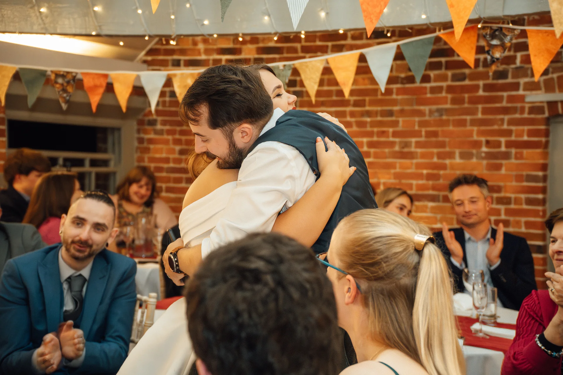 A groom enthusiastically embraces his bride in front of cheering guests during the wedding breakfast in a rustic brick venue decorated with bunting.