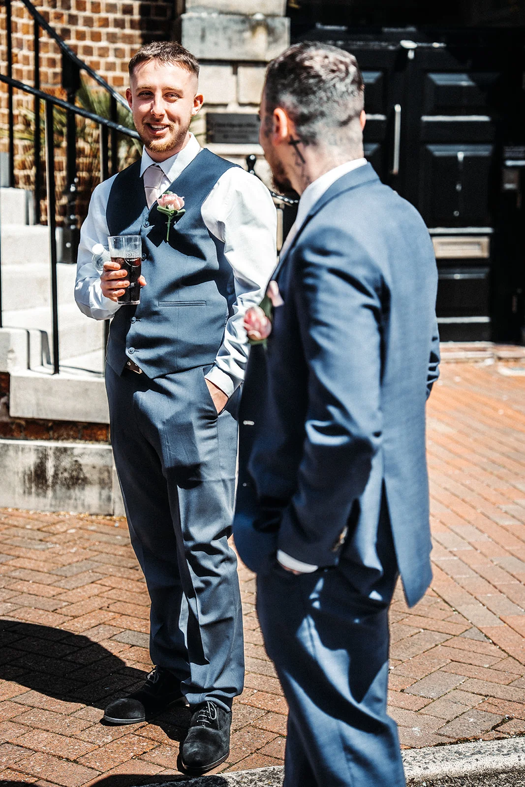 Two groomsmen in blue suits stand chatting outdoors in a sunny cobbled courtyard, with one smiling warmly while holding a drink.