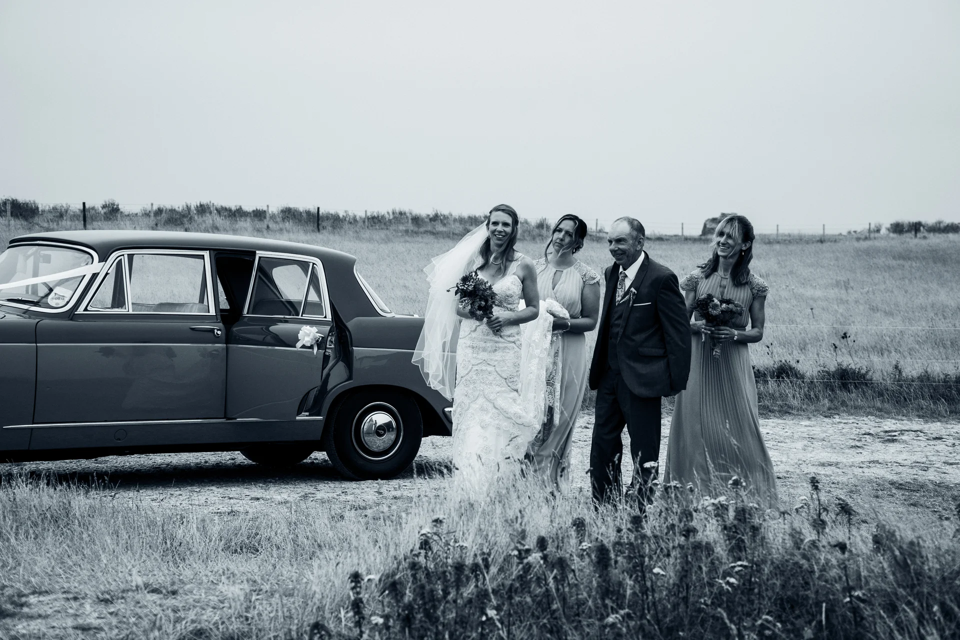 A bride with a veil and bouquet walks alongside her father and bridesmaids next to a classic vintage car parked in a field.