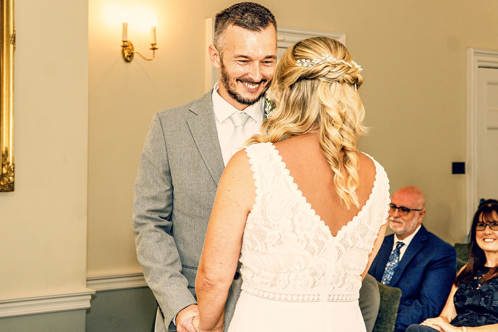 A groom in a grey suit smiles warmly at his bride during the ceremony while guests watch on lovingly from the background.