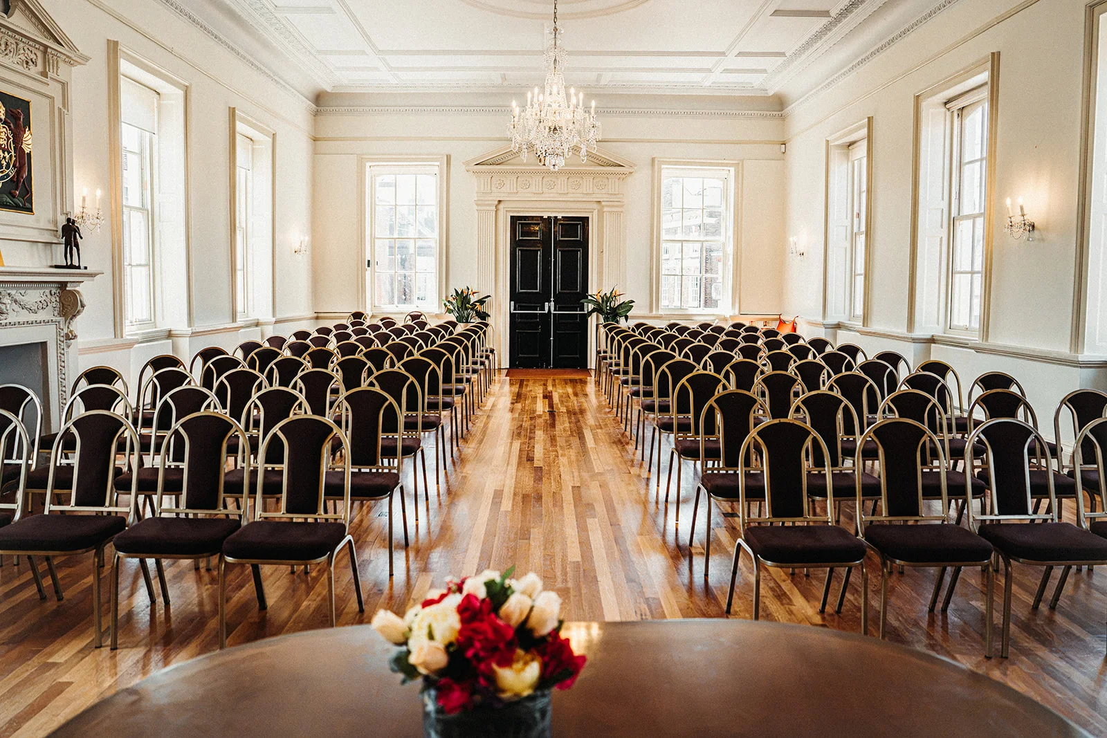 An elegant, light-filled Georgian manor ceremony room is arranged with rows of dark-chaired seating facing a central table adorned with a vibrant floral arrangement.