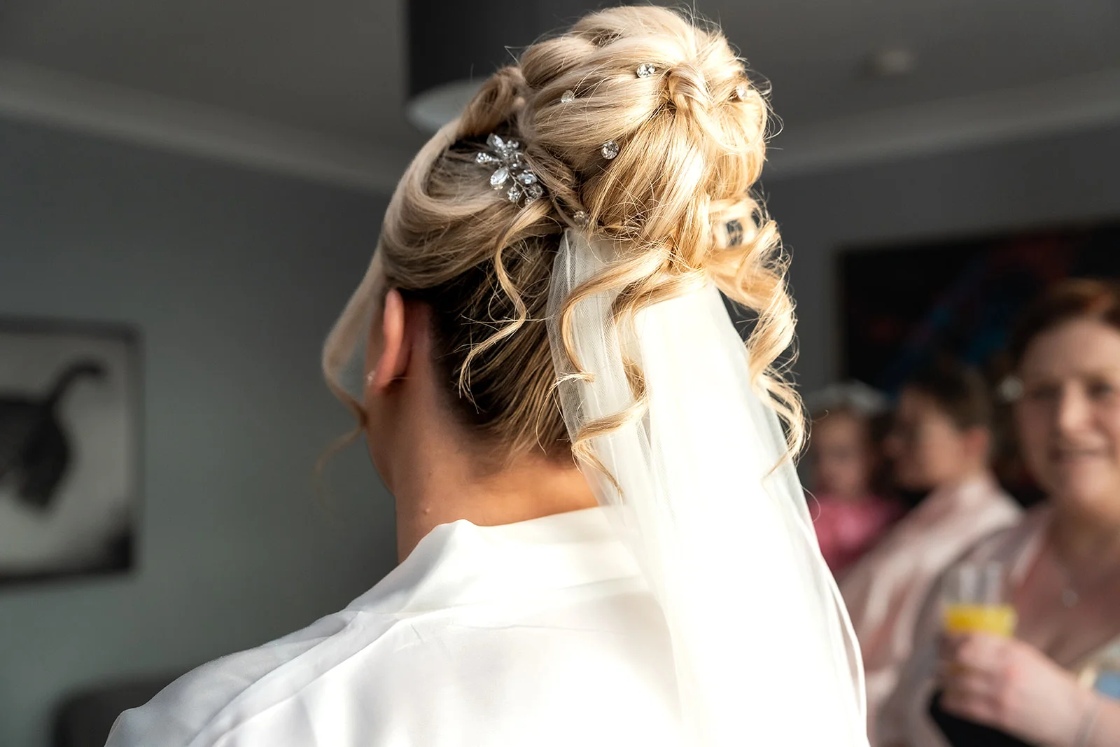 A blonde bride wearing a white getting ready robe showcases an intricate, textured updo adorned with sparkling hair pins and a delicate veil during bridal prep.