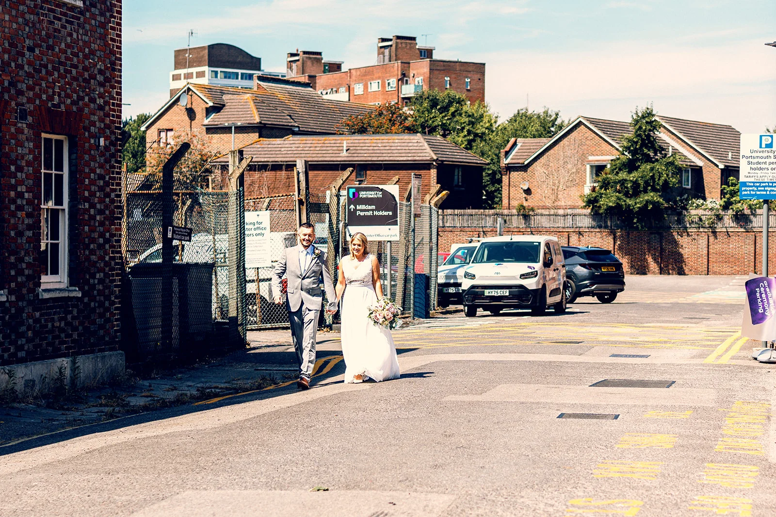 A bride and groom walk hand in hand down a sunlit city street, flanked by brick buildings and parked cars, with the bride carrying a delicate bouquet.