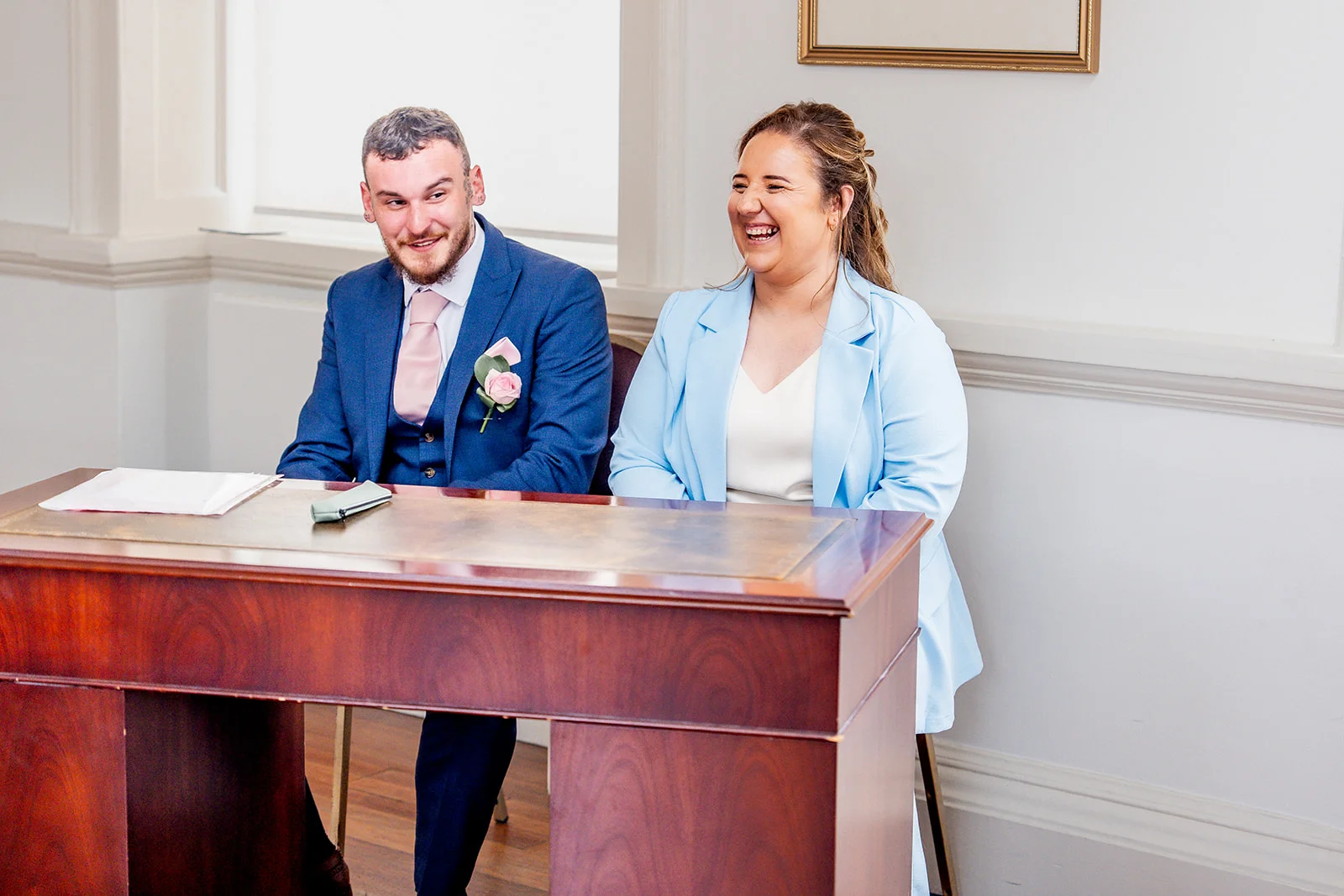 A newly married couple laughing joyfully while seated at a wooden desk during their civil ceremony in a registry office.