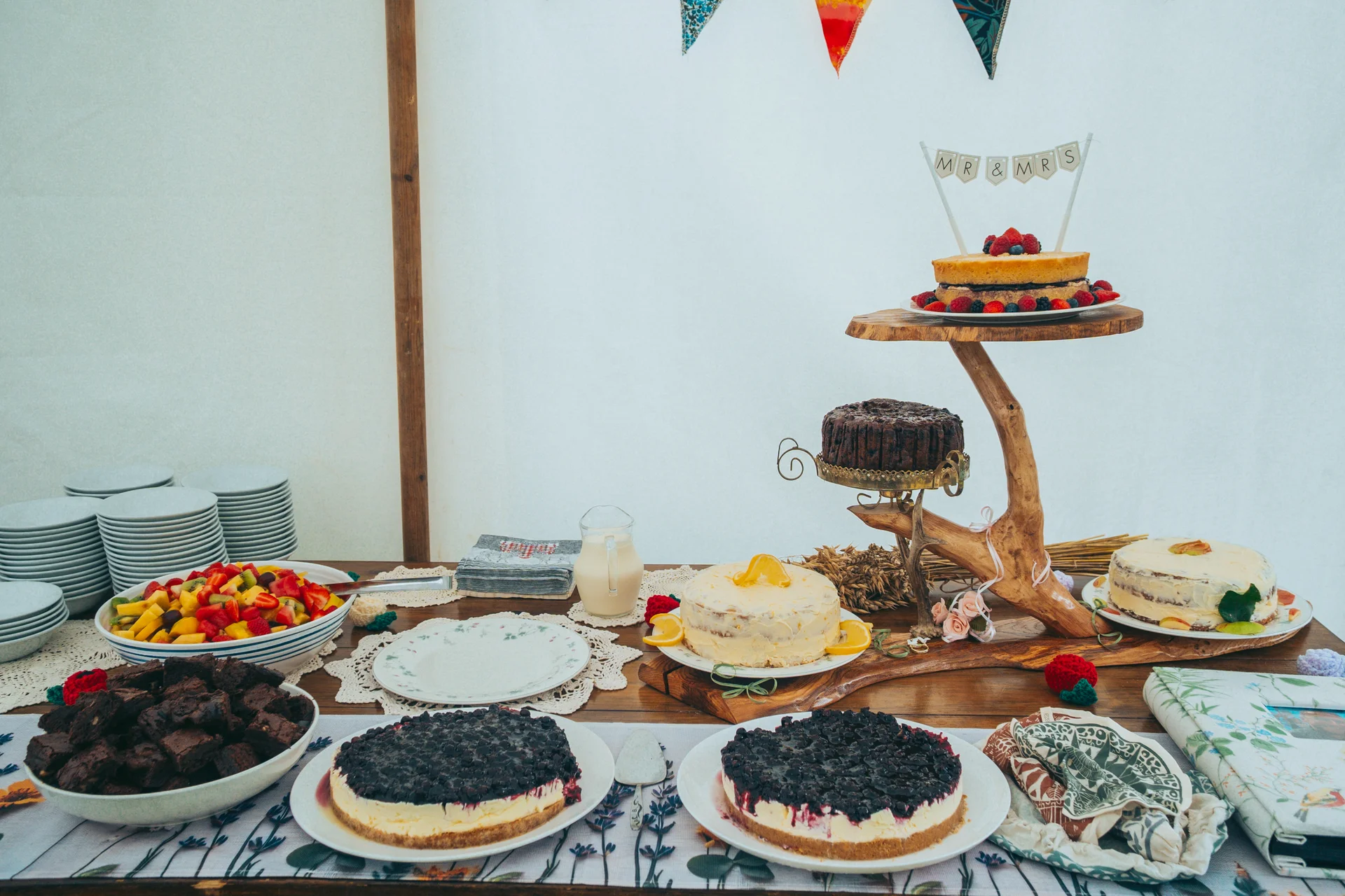 A rustic dessert table inside a marquee featuring various cakes, cheesecakes, brownies, and a fresh fruit bowl set up for a wedding reception.