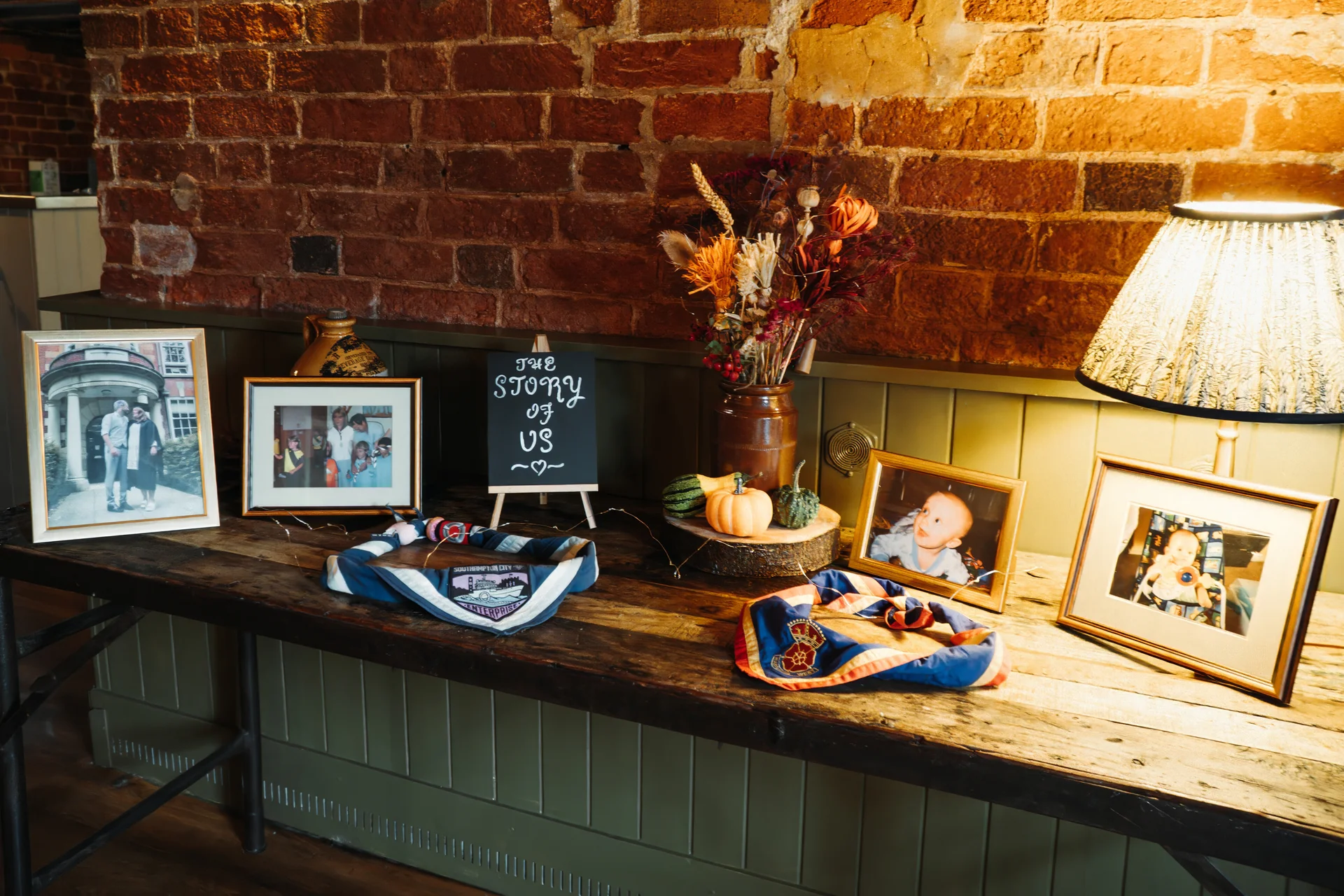 A rustic wooden table in a pub displays framed childhood photographs, a small chalkboard sign reading the story of us, and autumnal dried flowers against an exposed brick wall.