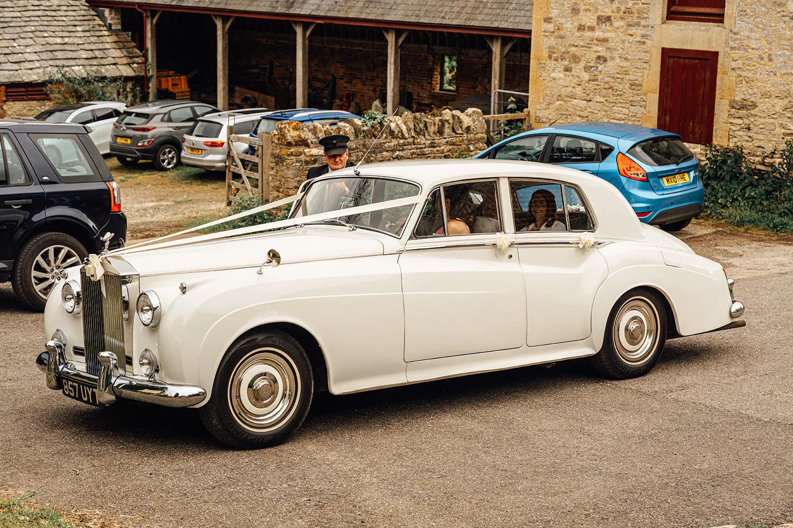 A classic white vintage wedding car adorned with white ribbons arrives outside a rustic stone barn venue with guests visible inside.