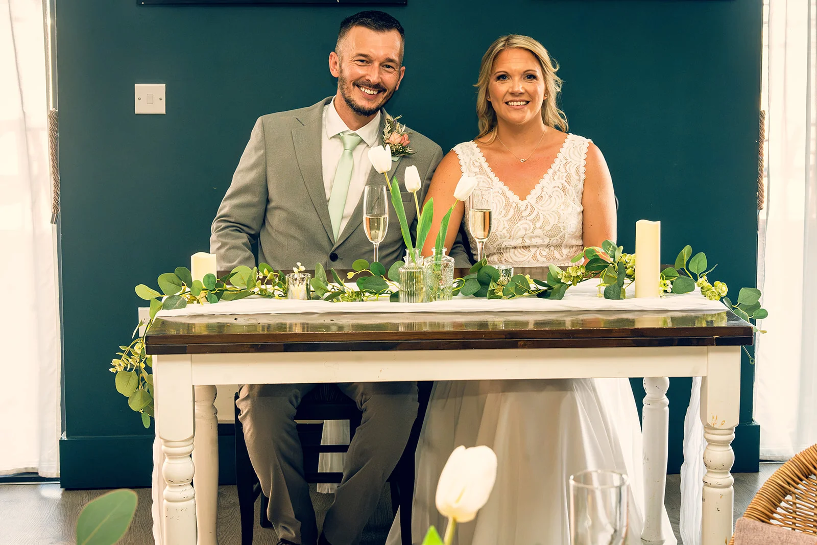 A smiling bride and groom sit together at a beautifully decorated top table featuring a greenery runner and white tulips against a dark teal wall.