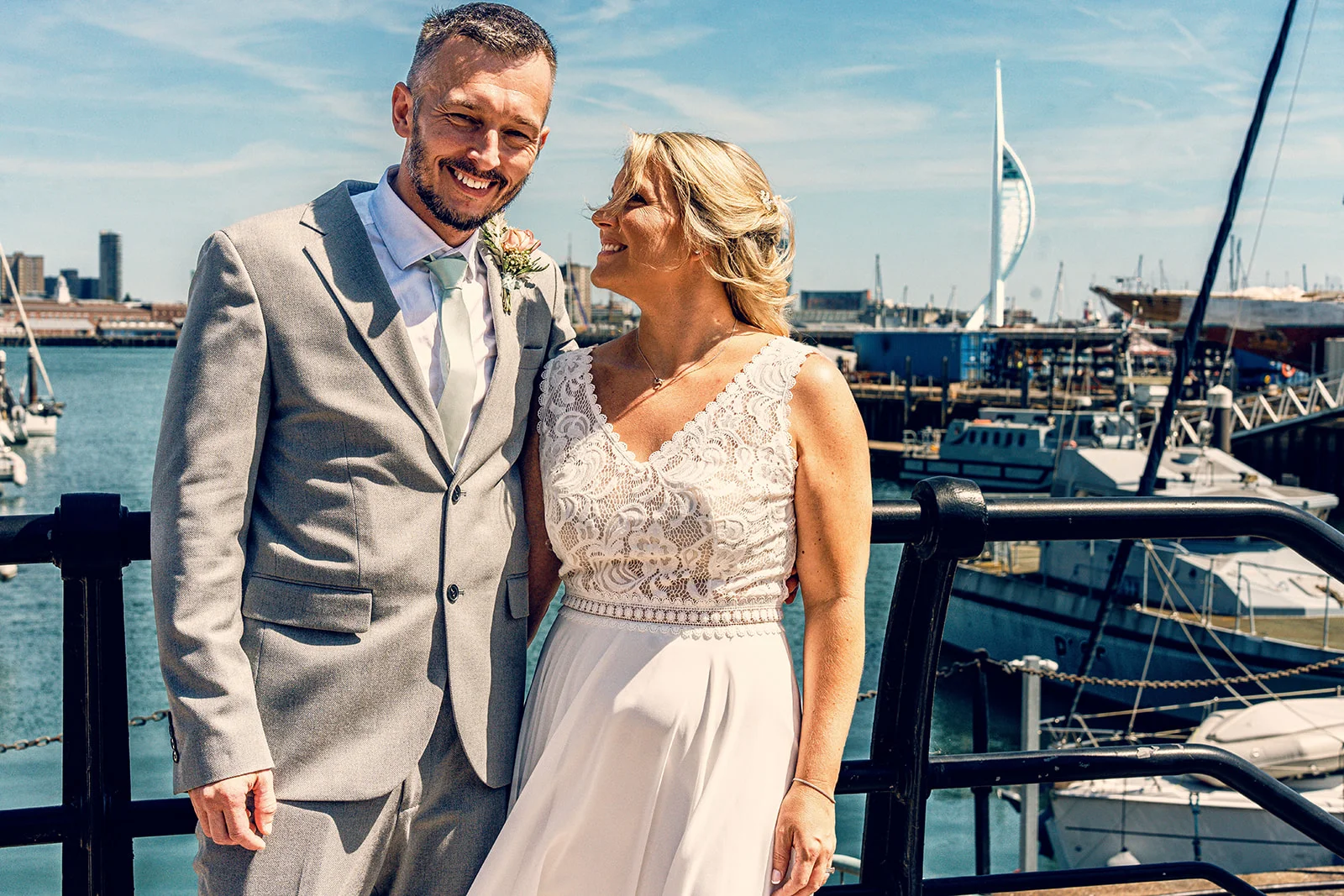 A smiling bride and groom share a loving glance during their sunny summer wedding by the harbour, with boats and a coastal backdrop.