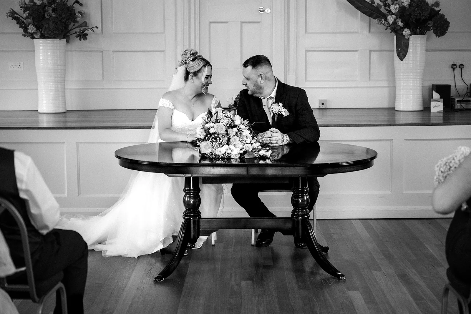A newly married couple lovingly gaze at each other while seated at a dark wooden table to sign their register during a civil ceremony.