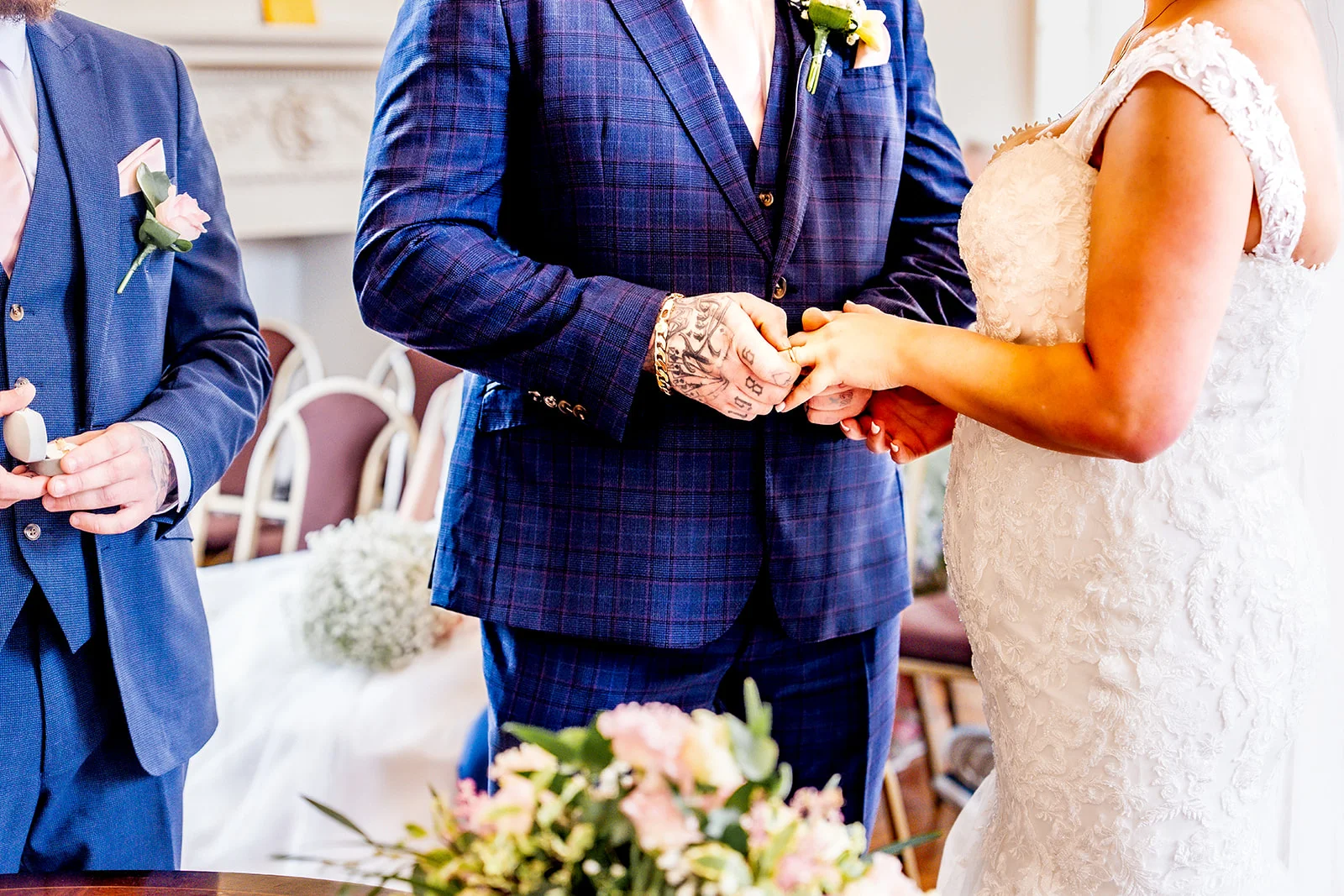 Close-up of a groom wearing a blue checked suit and the bride exchanging rings during the wedding ceremony, with a groomsman holding the ring box nearby.