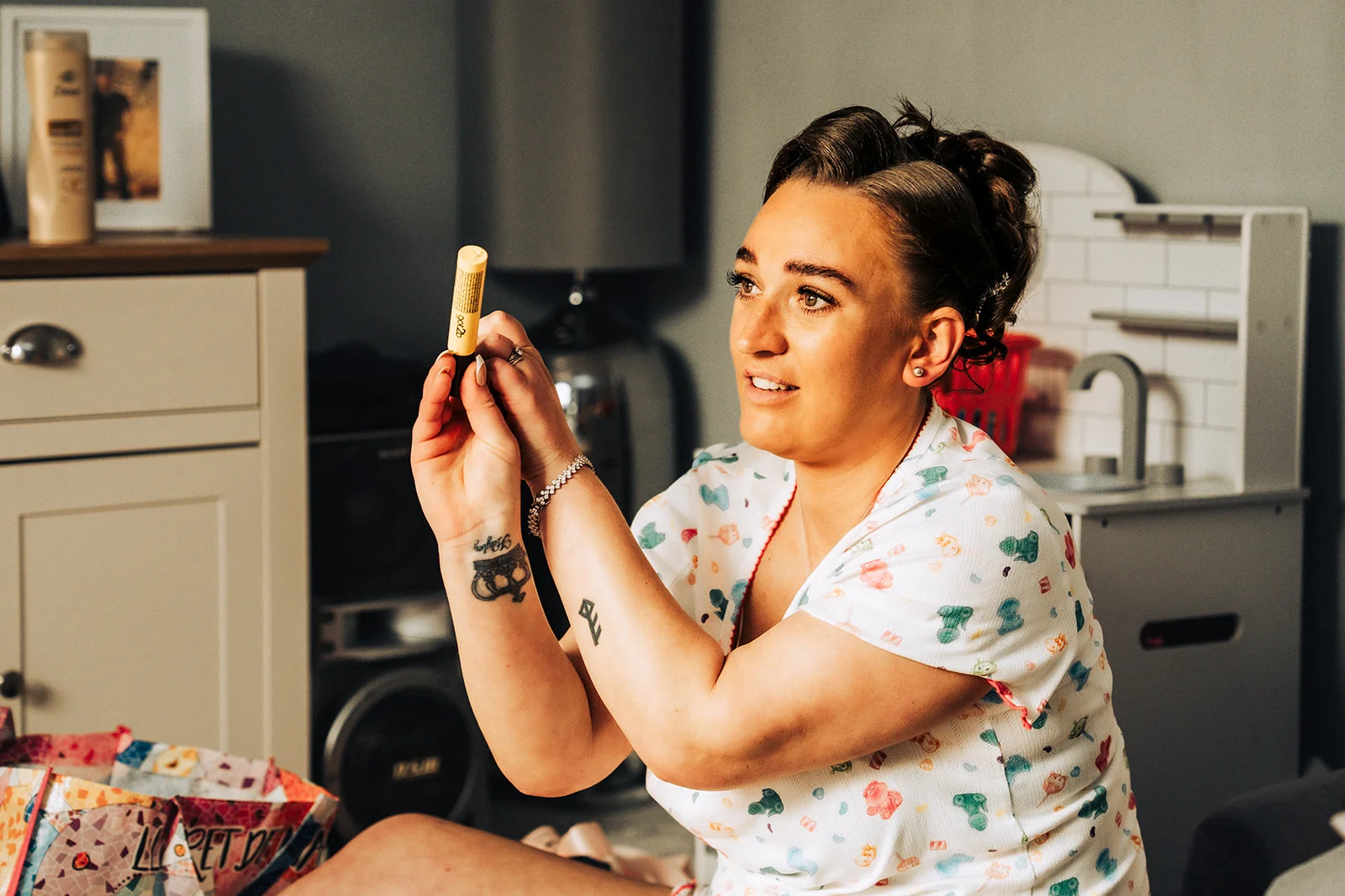 A bride in patterned pyjamas holds up a small yellow container while getting ready in a brightly lit room.