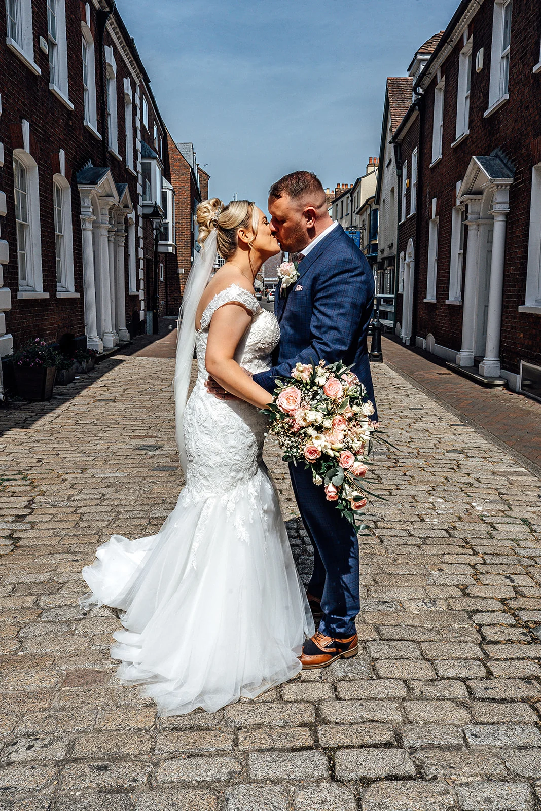 A bride and groom share a tender kiss while standing on a historic cobbled street lined with traditional brick buildings on a bright sunny day.