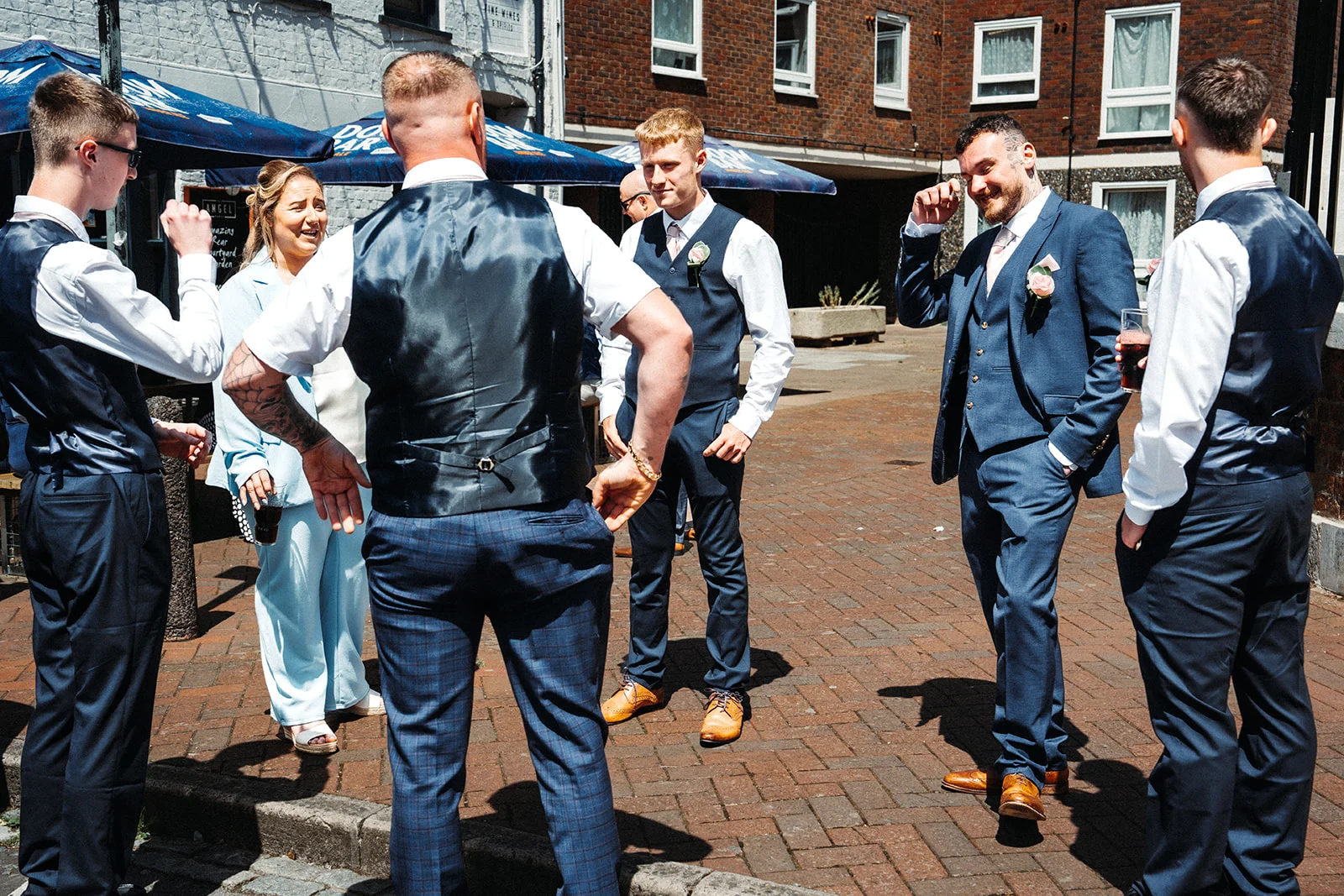 A group of groomsmen in navy blue suits and waistcoats chat and laugh together in a sunlit pub courtyard.