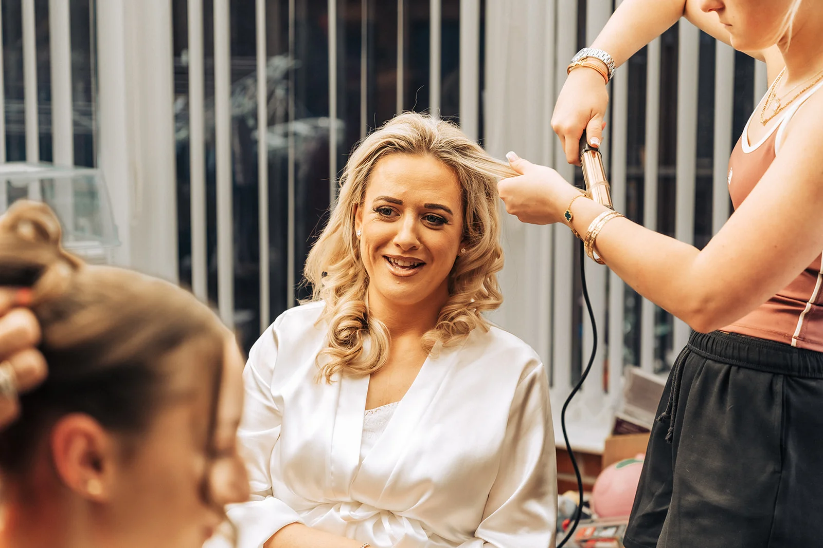 A smiling bride wearing a white satin robe has her blonde hair styled with a curling iron by a stylist during wedding morning preparations.