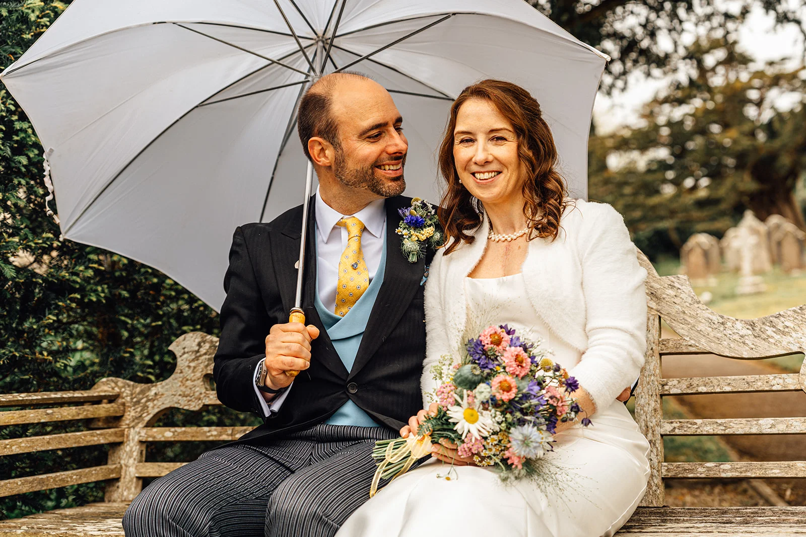 A newlywed couple sits smiling together on an outdoor bench holding a large clear umbrella, with the bride holding a colourful wild flower bouquet.