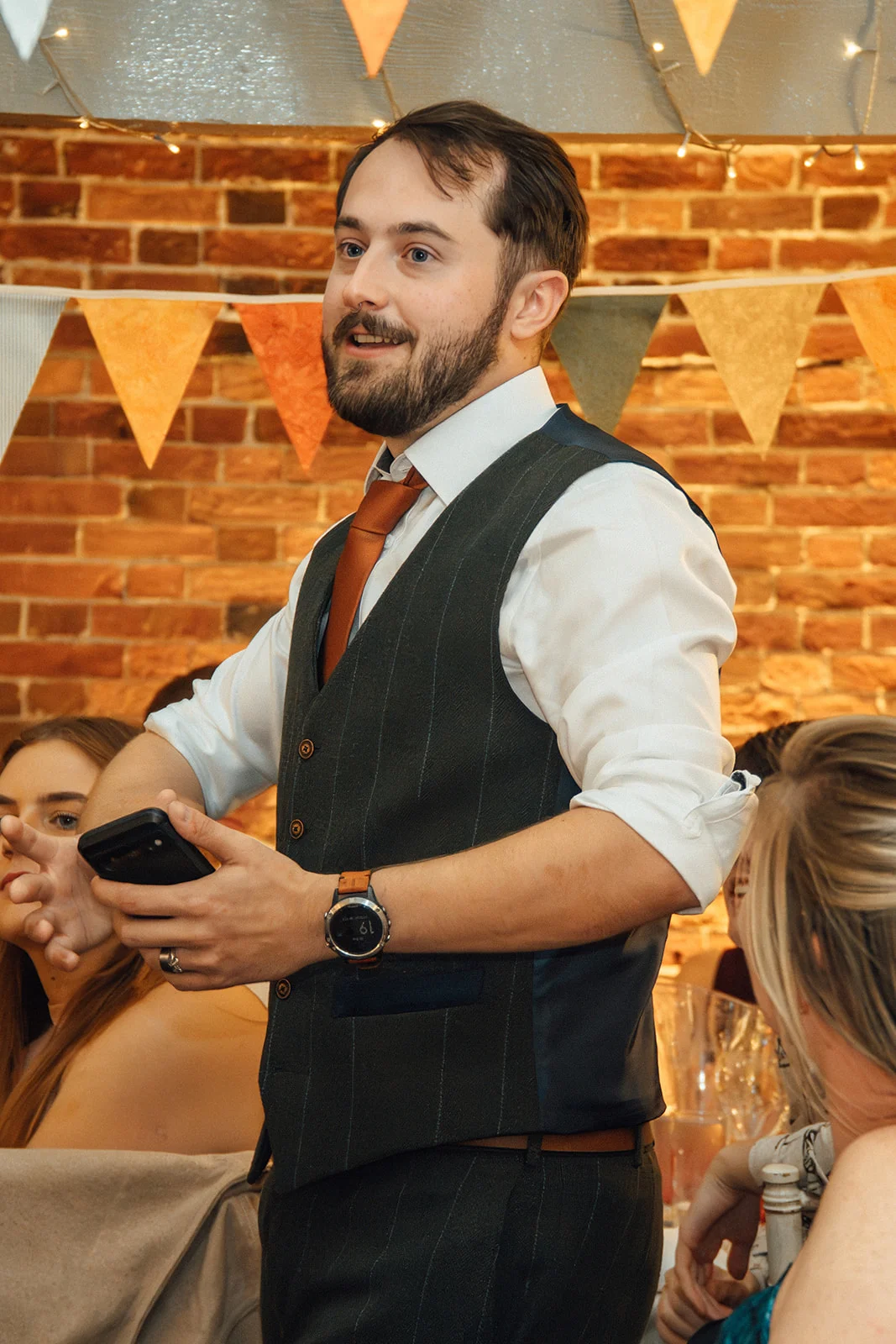 A smiling man gives a speech holding a phone in front of an exposed brick wall decorated with bunting and fairy lights.