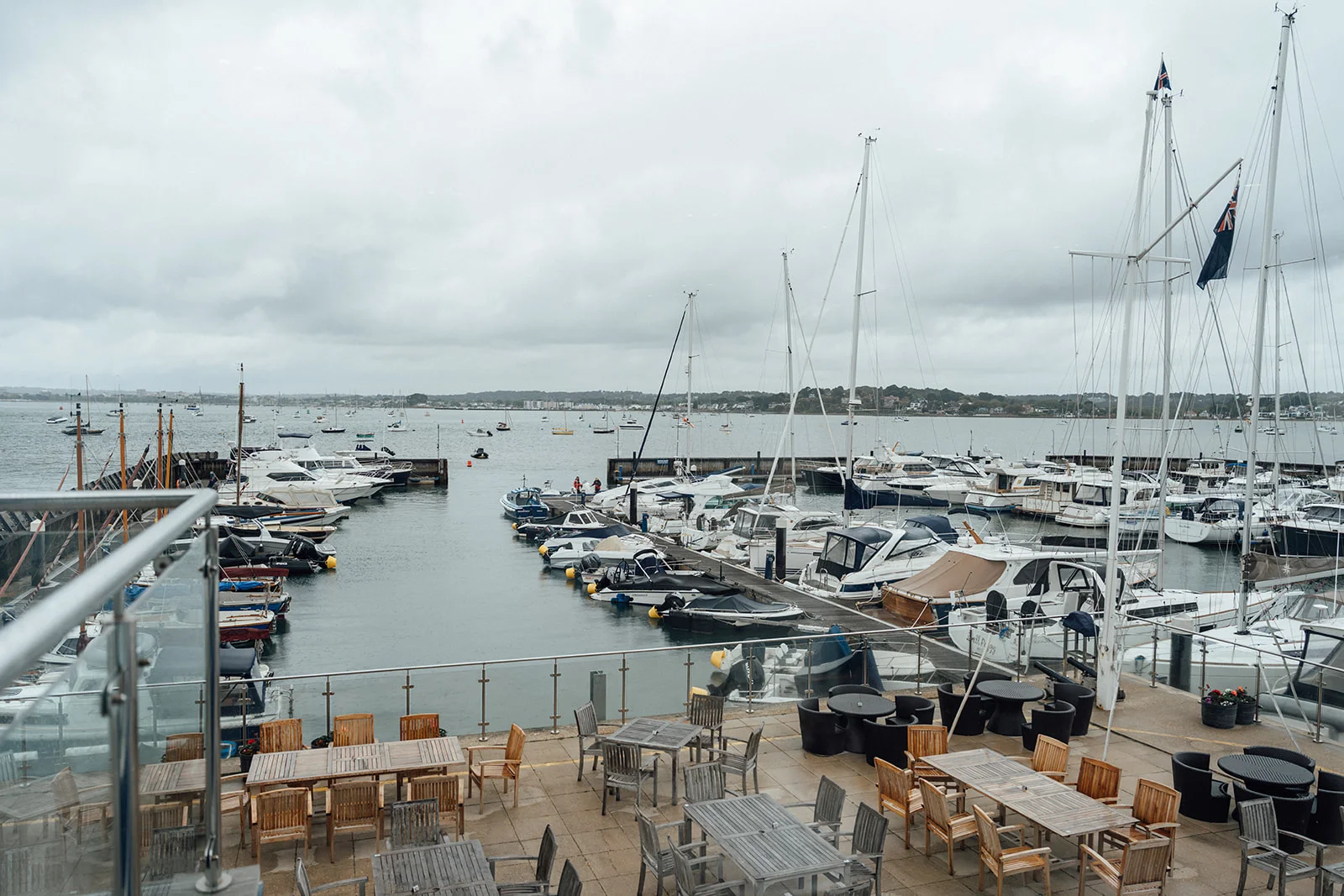 An elevated view overlooking a busy marina filled with numerous moored boats and yachts under an overcast grey sky, with outdoor dining tables arranged on a paved terrace in the foreground.