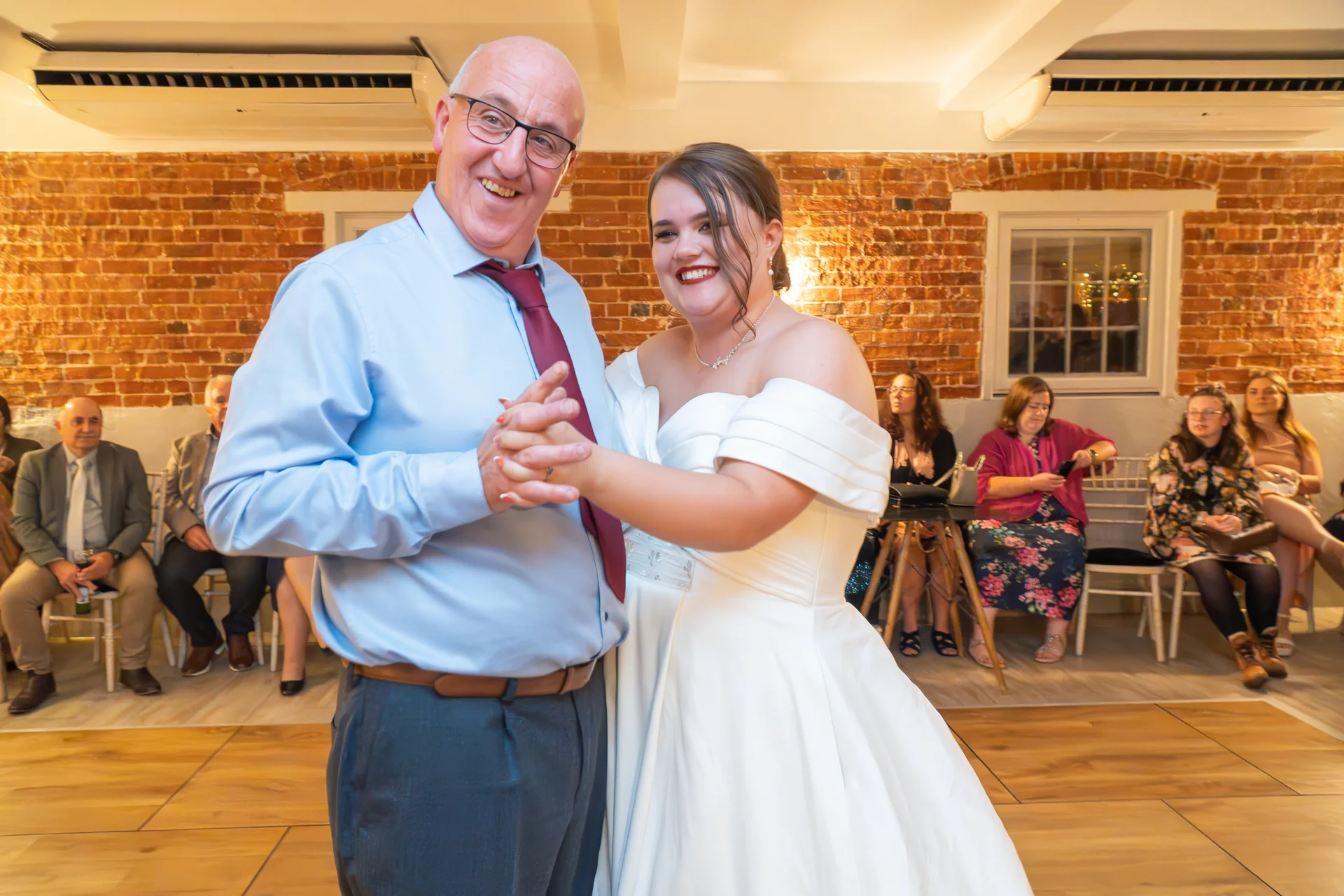 A smiling bride shares a joyful father-daughter dance on a wooden dance floor in front of an exposed brick wall.