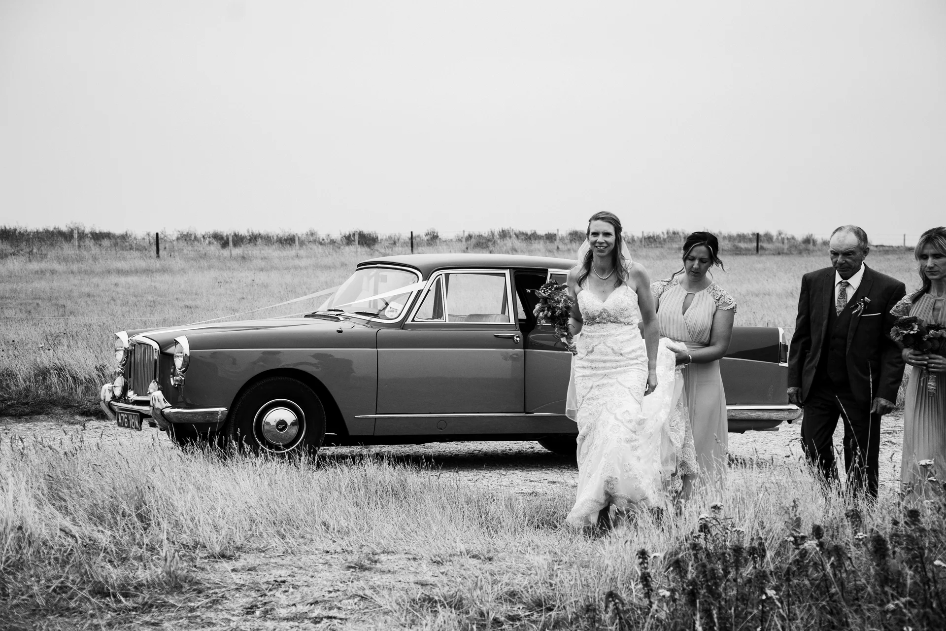 A bride in a lace wedding dress and veil steps out of a vintage car in a rural landscape, accompanied by her bridesmaid and father.