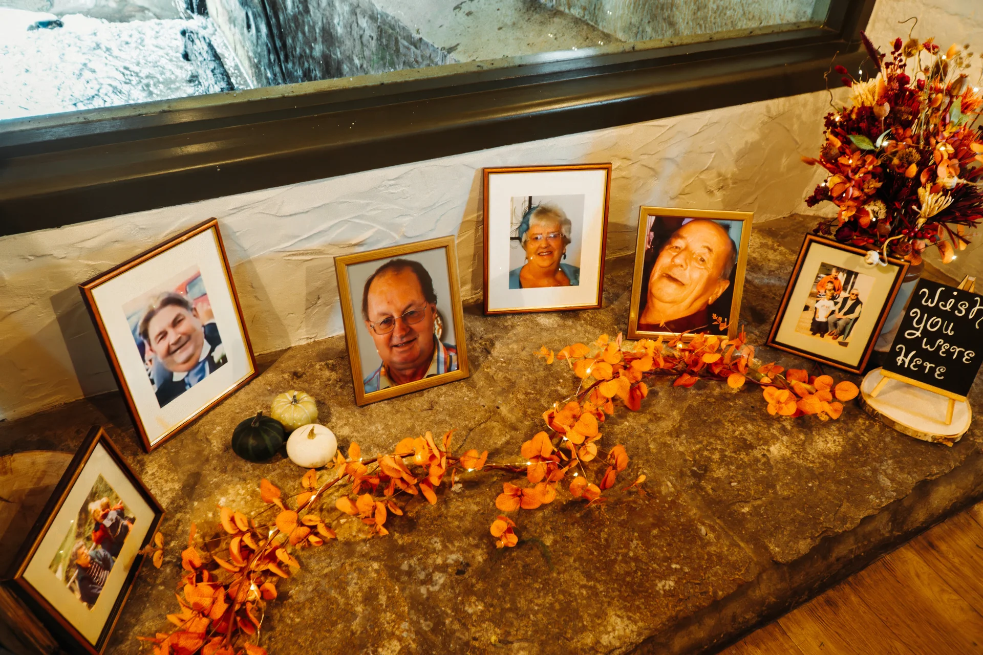 A thoughtful memory table features framed photographs of family members decorated with autumn leaves, mini pumpkins, fairy lights, and a chalkboard sign.